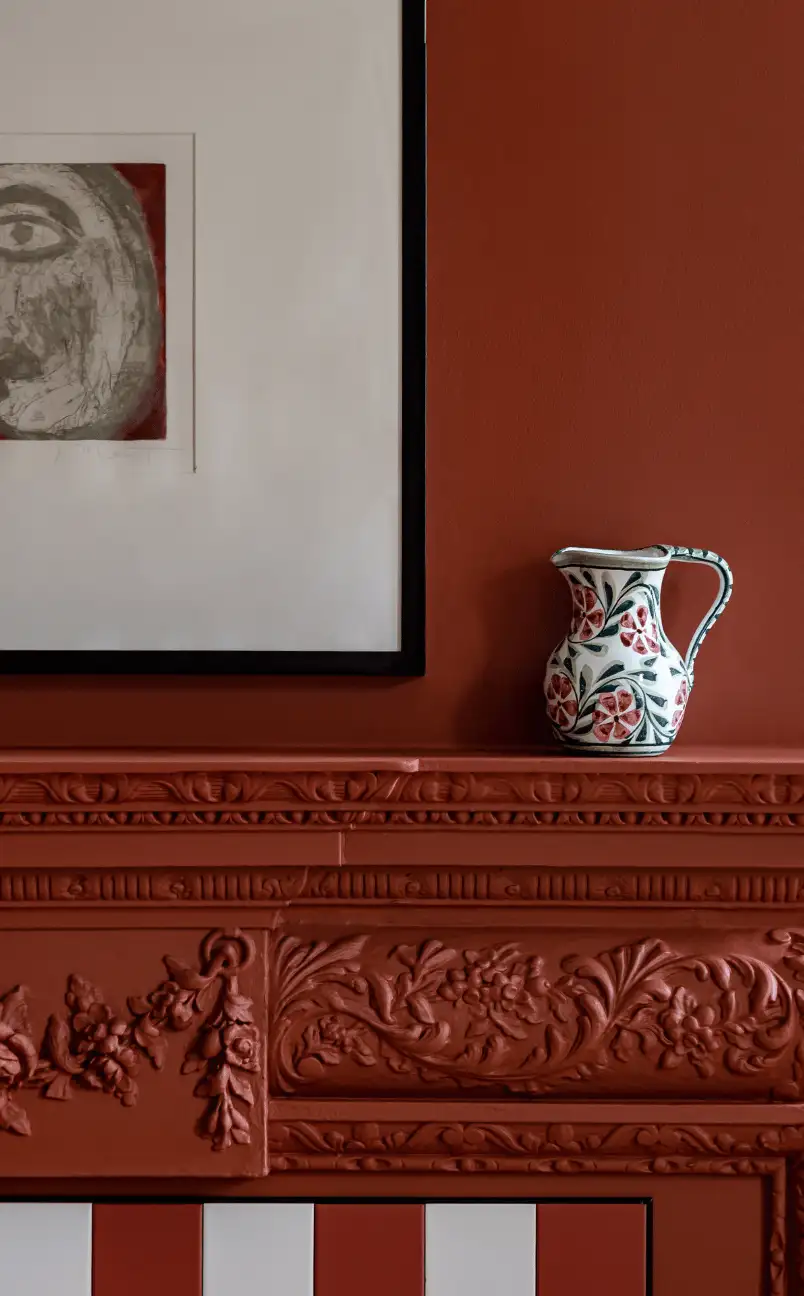 Ceramic pitcher with floral design placed on a detailed terracotta-colored mantelpiece beneath a framed abstract face artwork.