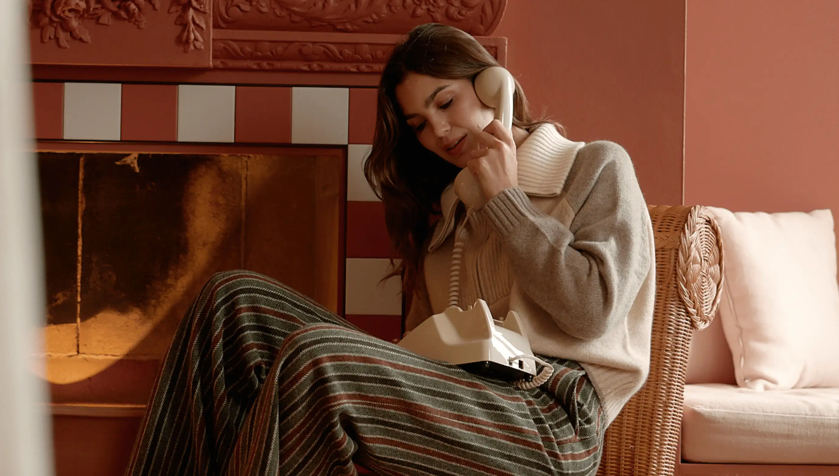 Woman sitting in a wicker chair near a fireplace, talking on a vintage beige corded telephone.