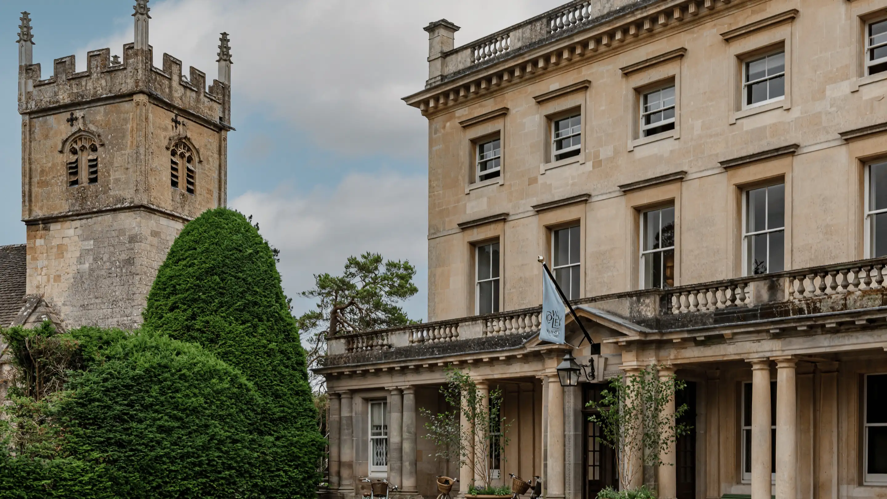 Stone building with classical columns and multiple windows next to a medieval-style stone tower and manicured green bushes.