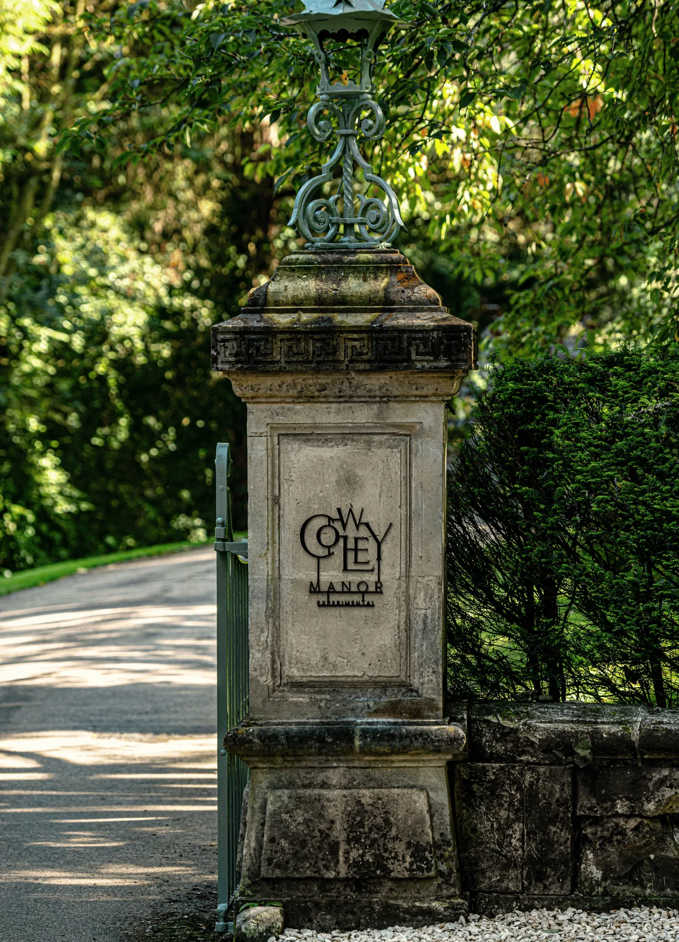 Stone gate pillar with decorative green ironwork and a sign that reads 'Cowley Manor' surrounded by greenery and a shaded driveway.