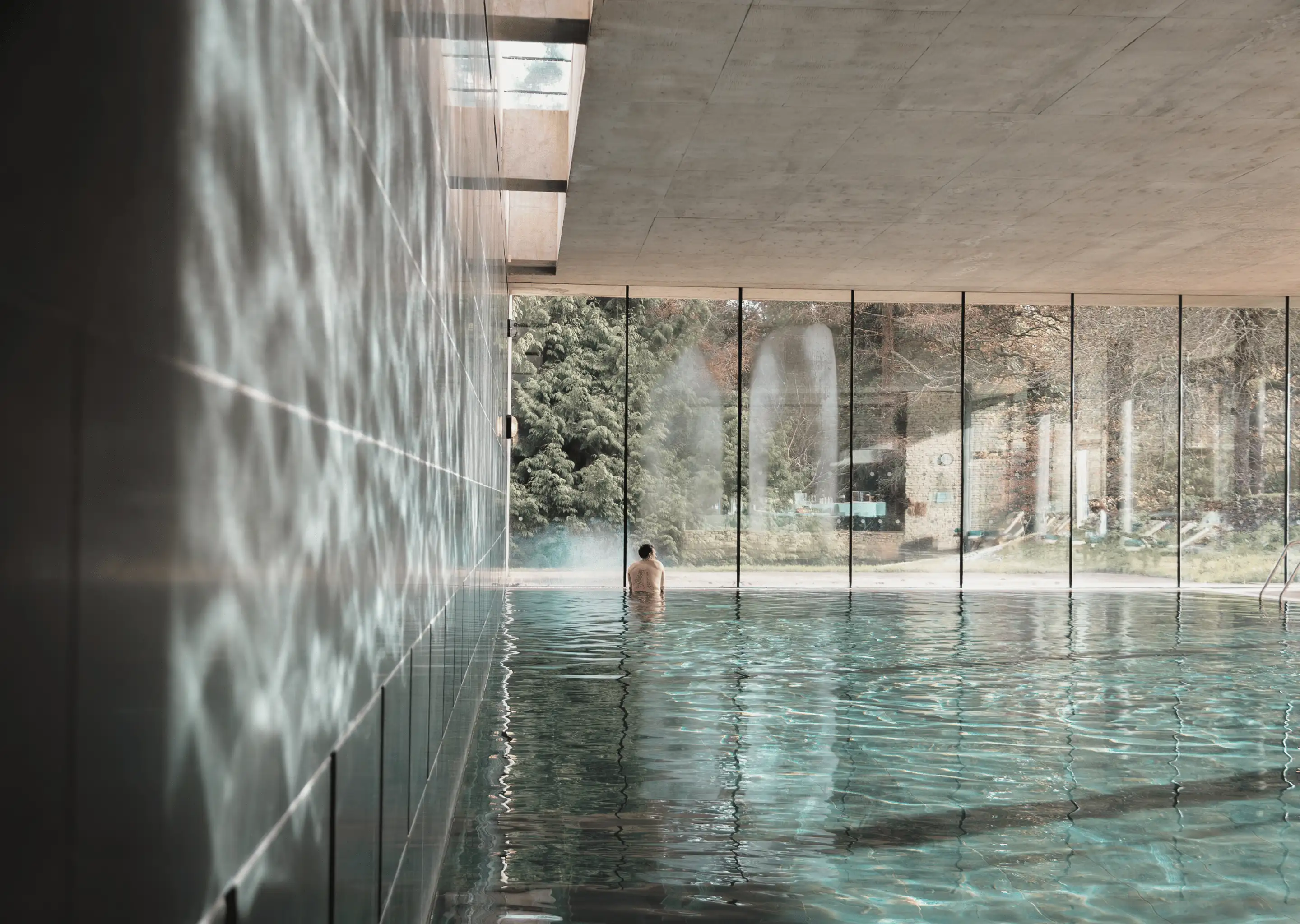 Indoor pool with a person sitting at the edge, large floor-to-ceiling windows showing trees outside.