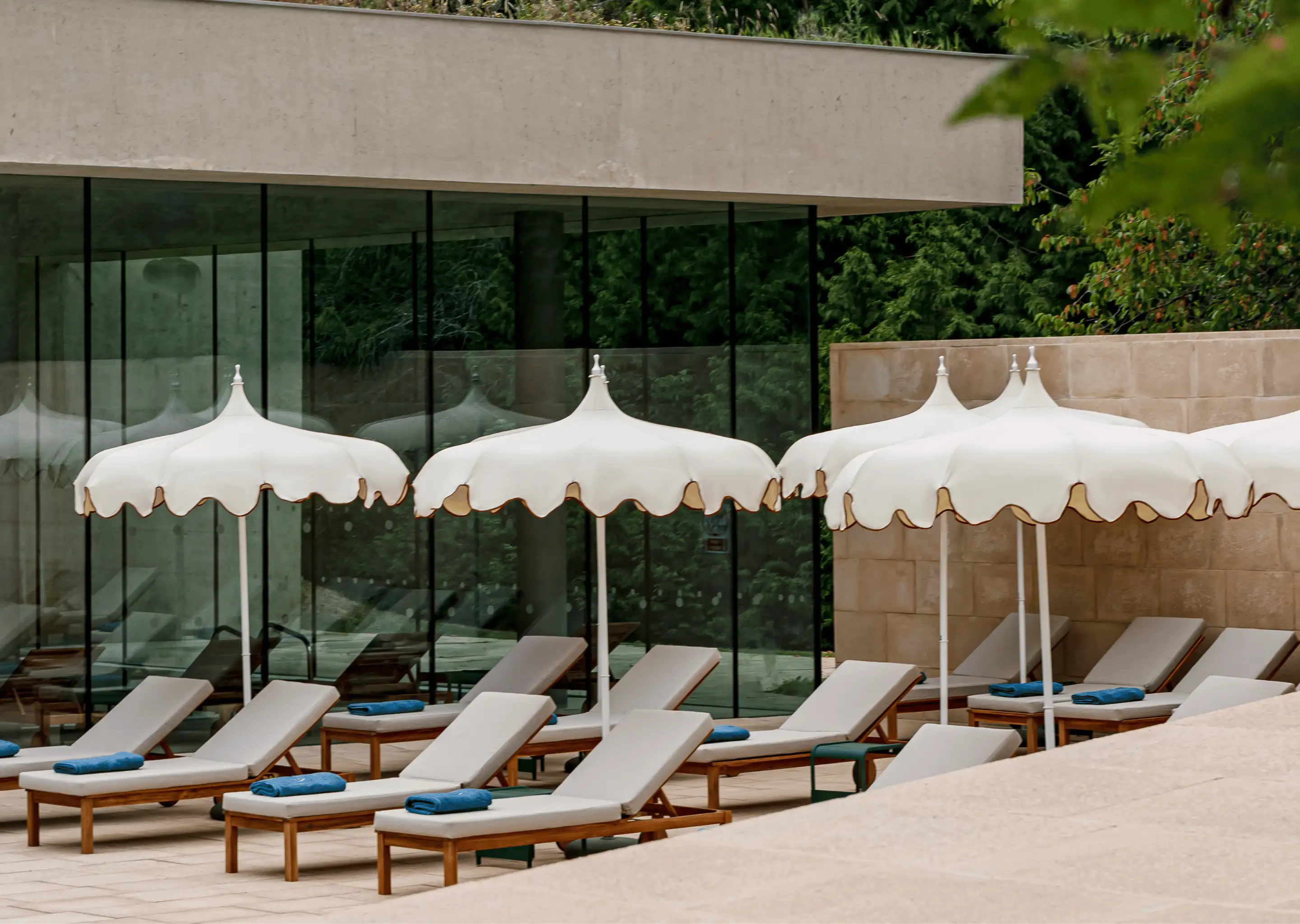 Outdoor poolside lounge area with beige cushioned wooden sunbeds and white scalloped umbrellas, each with a blue towel.
