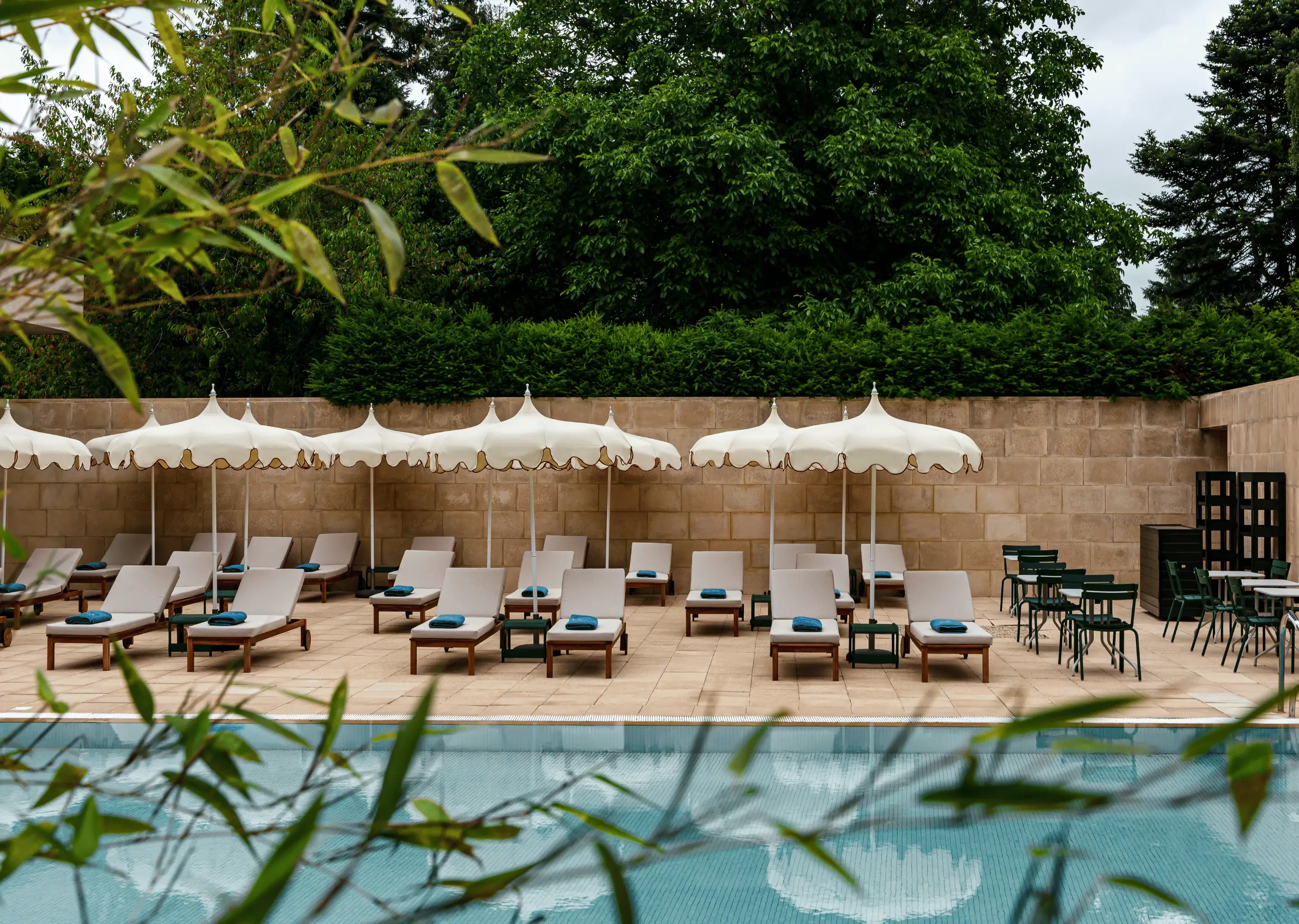 Poolside area with multiple beige lounge chairs each topped with a folded blue towel, white umbrellas, and green chairs and tables against a stone wall with trees in the background.