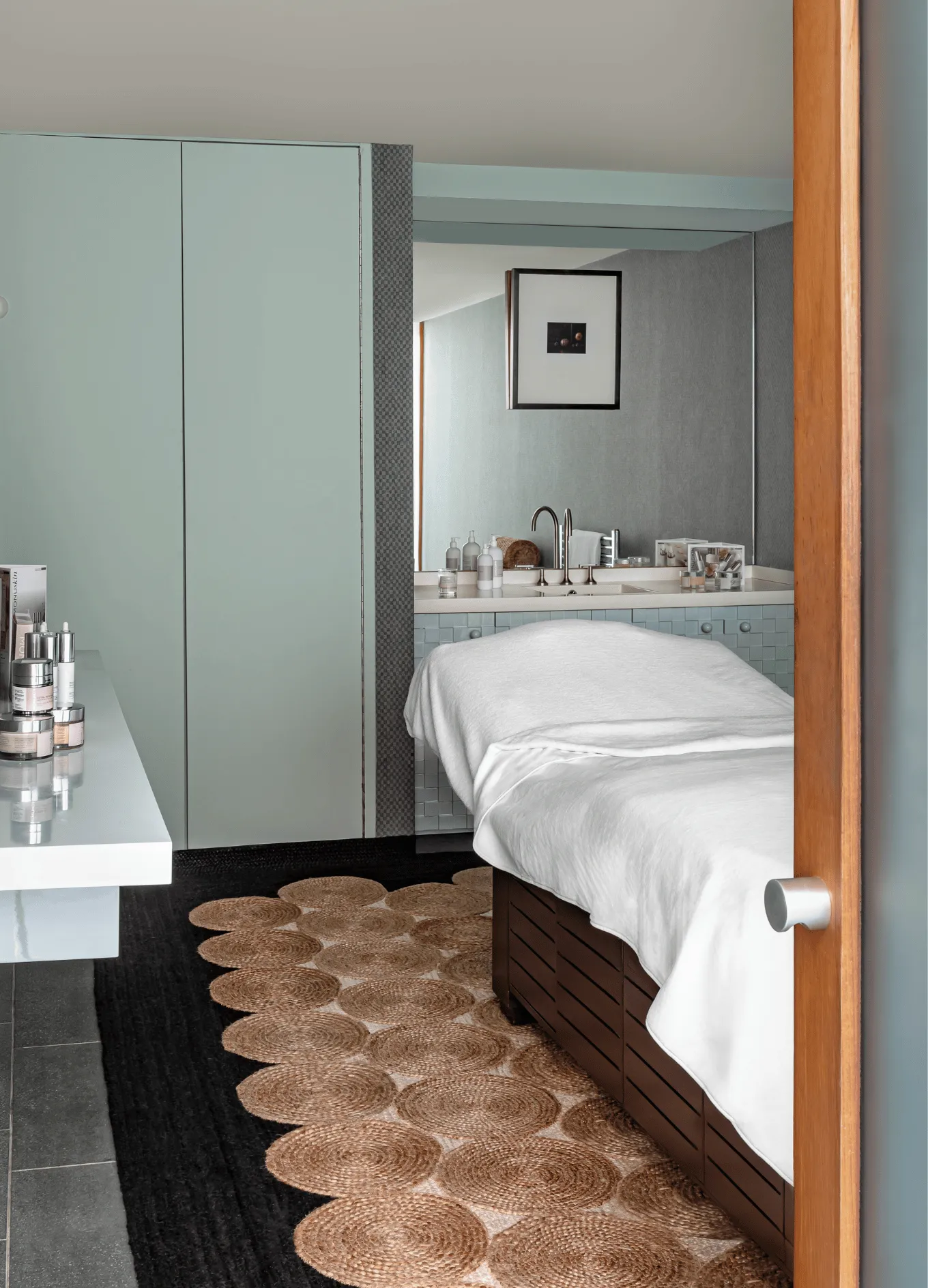 Massage table covered with a white towel in a modern wellness room with skincare products on a countertop and a large mirror.