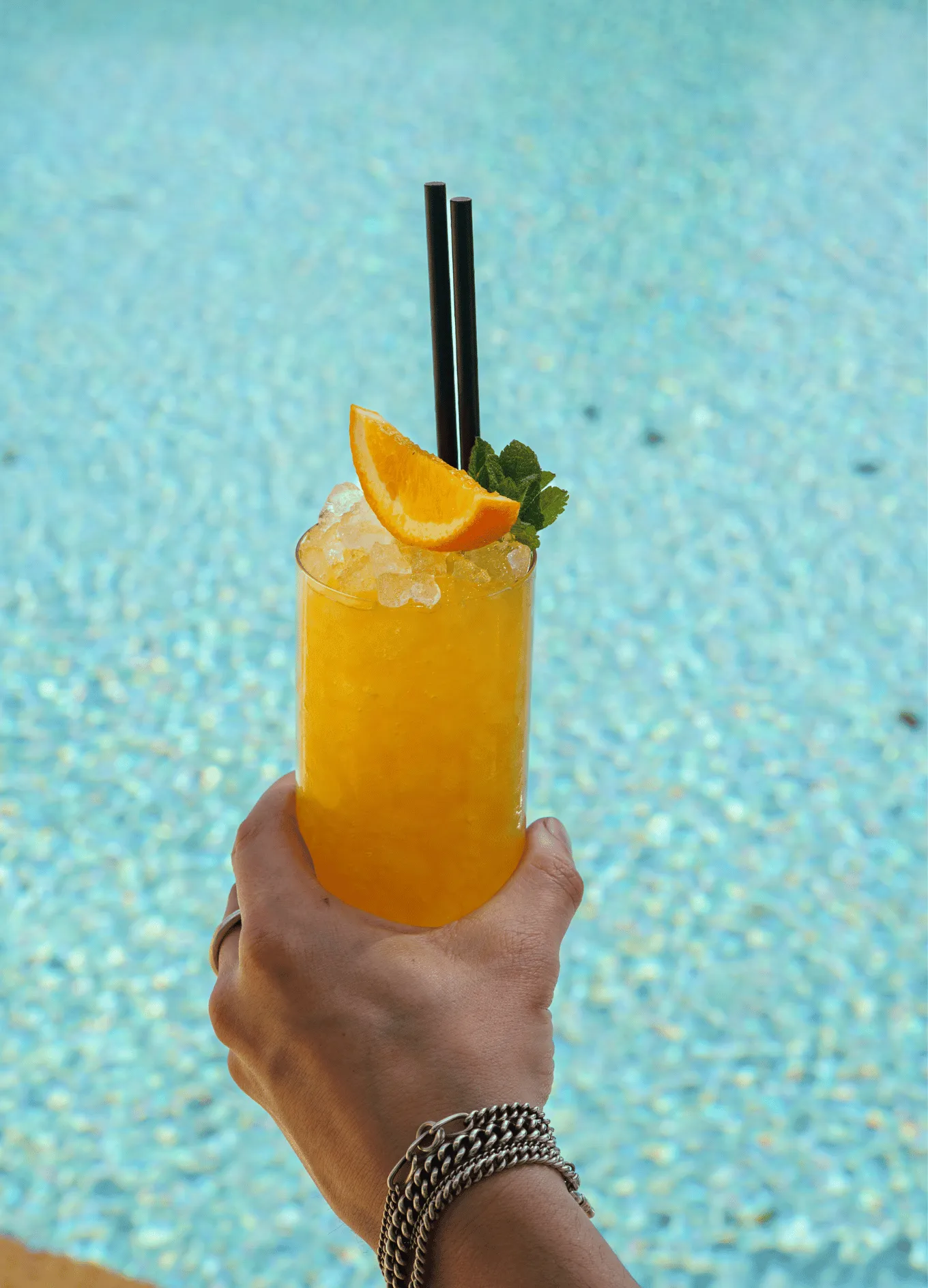 Hand holding a yellow iced cocktail garnished with an orange slice and mint leaves, with a swimming pool in the background.