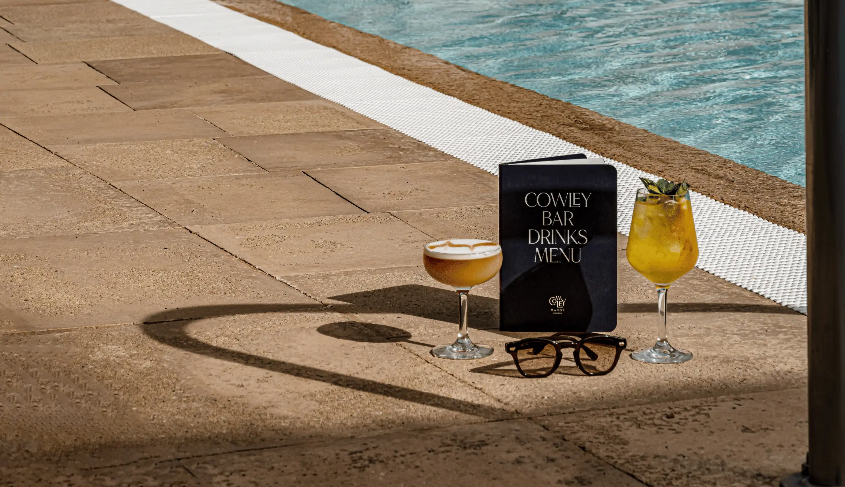 Two cocktails, a pair of eyeglasses, and a Cowley Bar drinks menu placed on the poolside pavement beside a swimming pool.