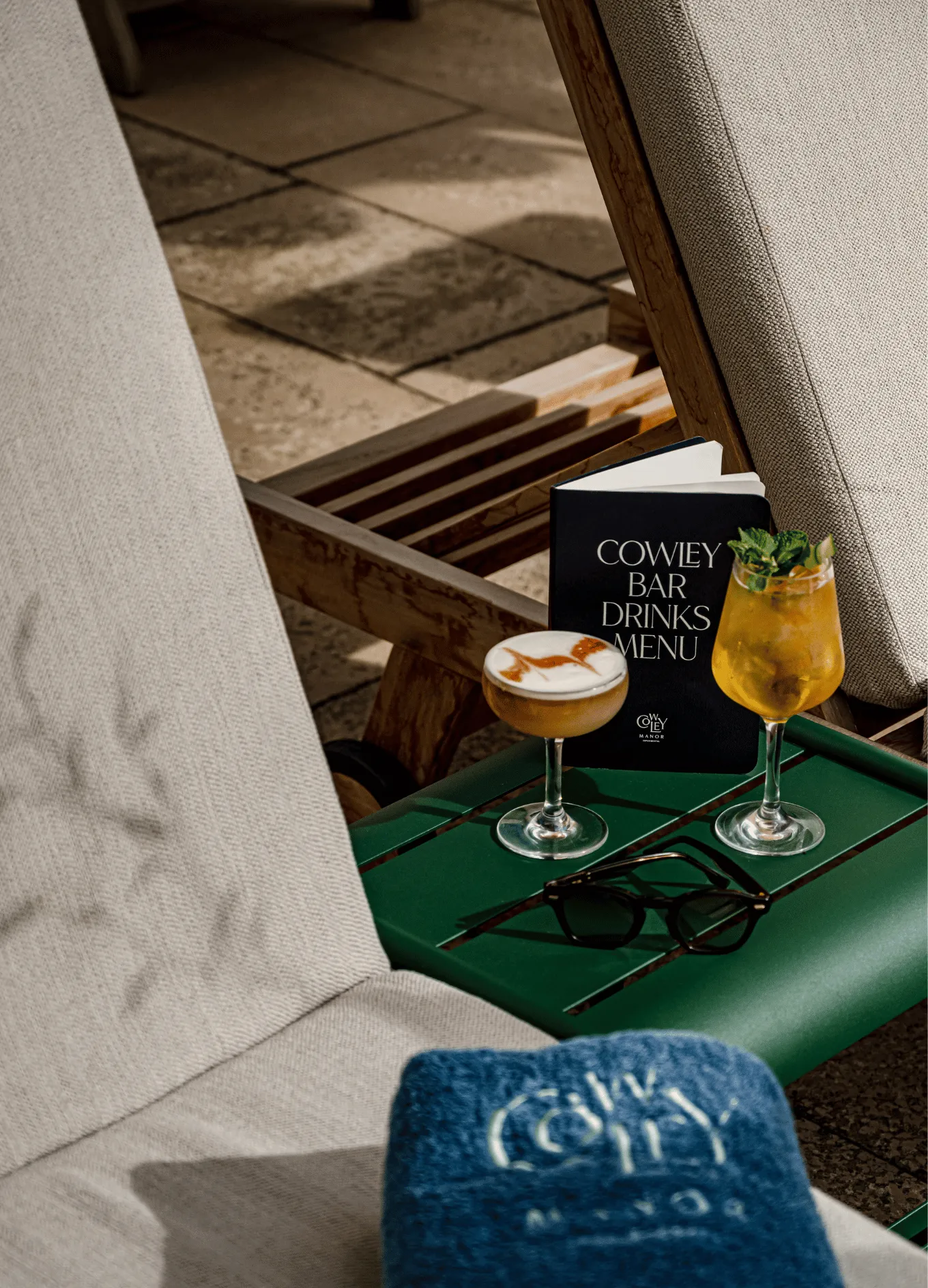 Two cocktails and a black Cowley Bar Drinks Menu on a green table between beige lounge chairs, with sunglasses and a blue Cowley Manor towel nearby.