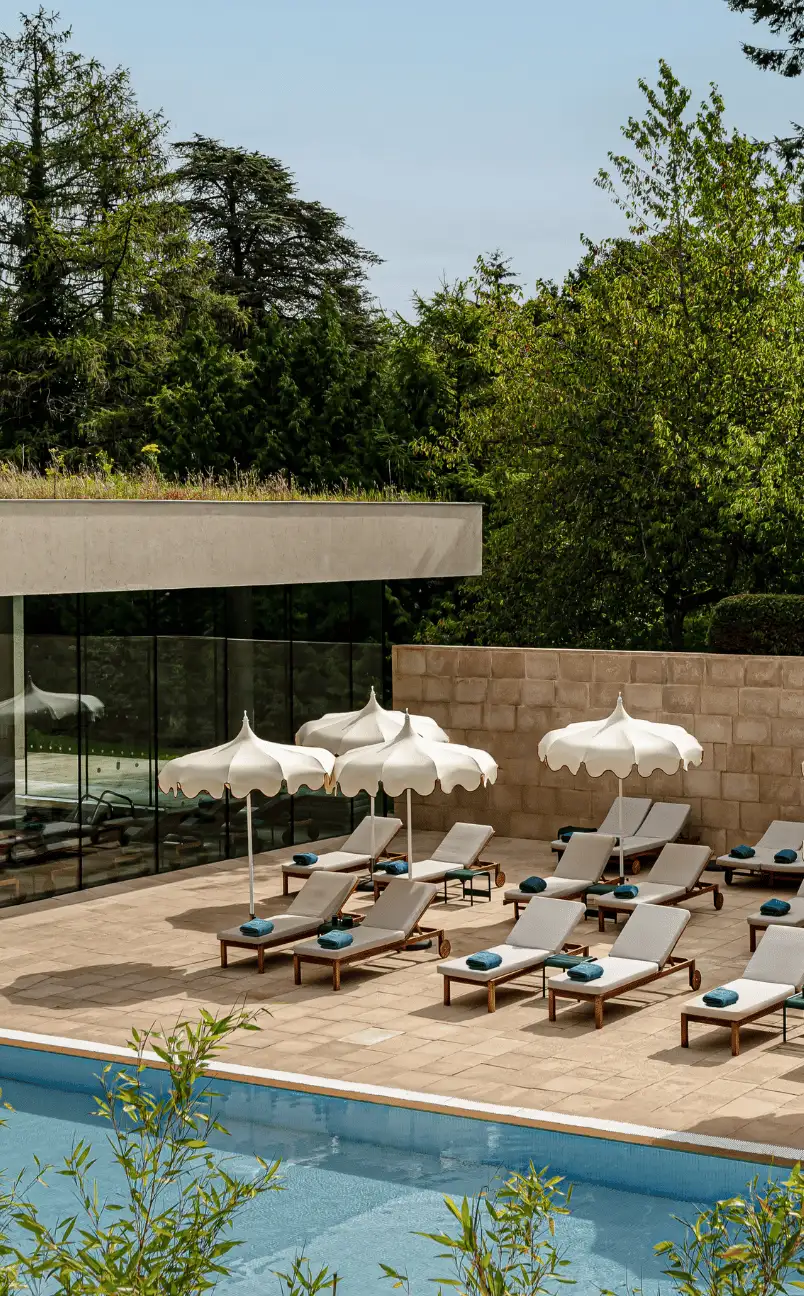 Outdoor pool area with white lounge chairs and umbrellas on a stone deck surrounded by trees and a glass building.