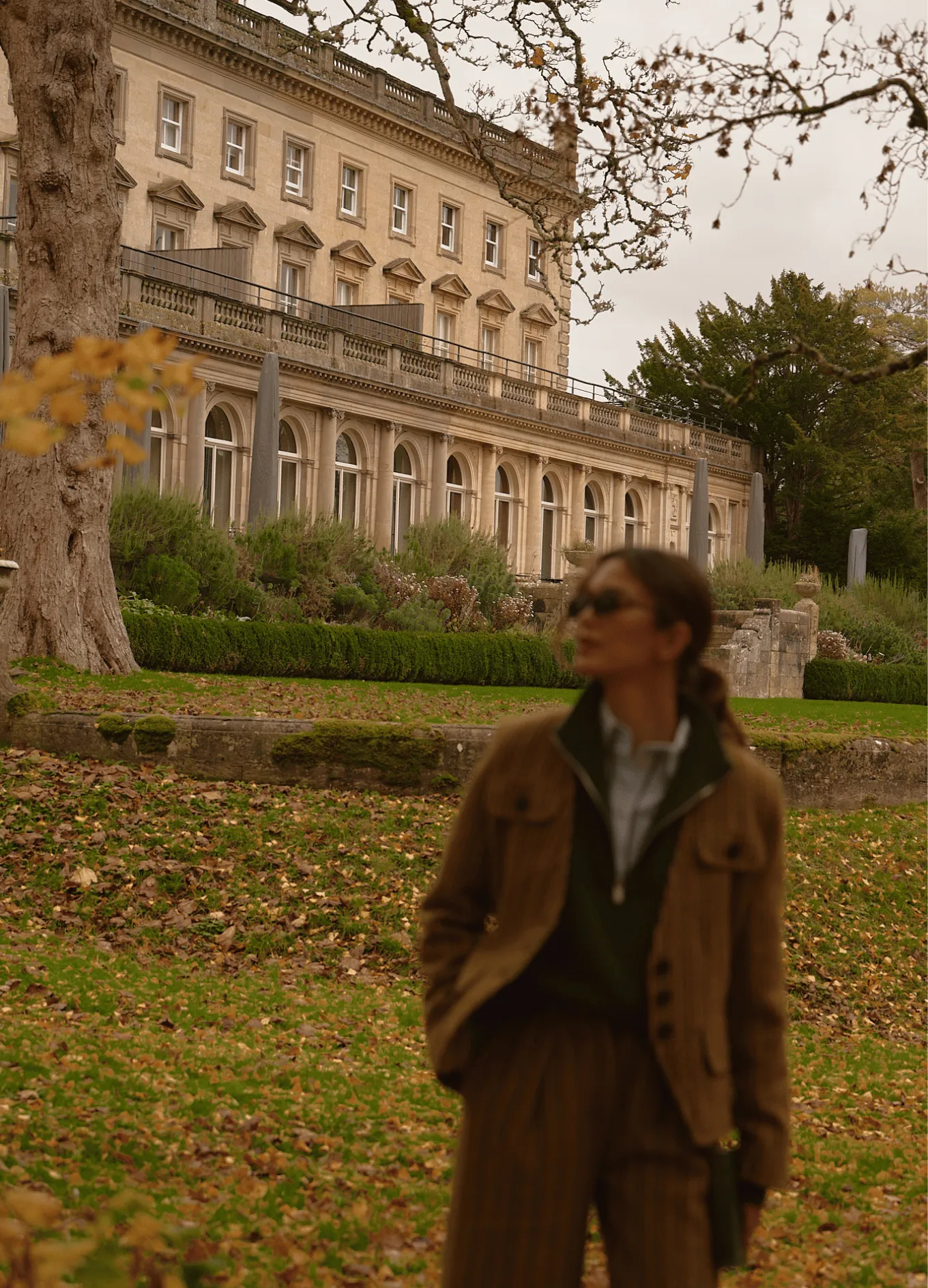 Woman wearing sunglasses and a brown jacket standing in front of a large historic building with arched windows and manicured gardens.
