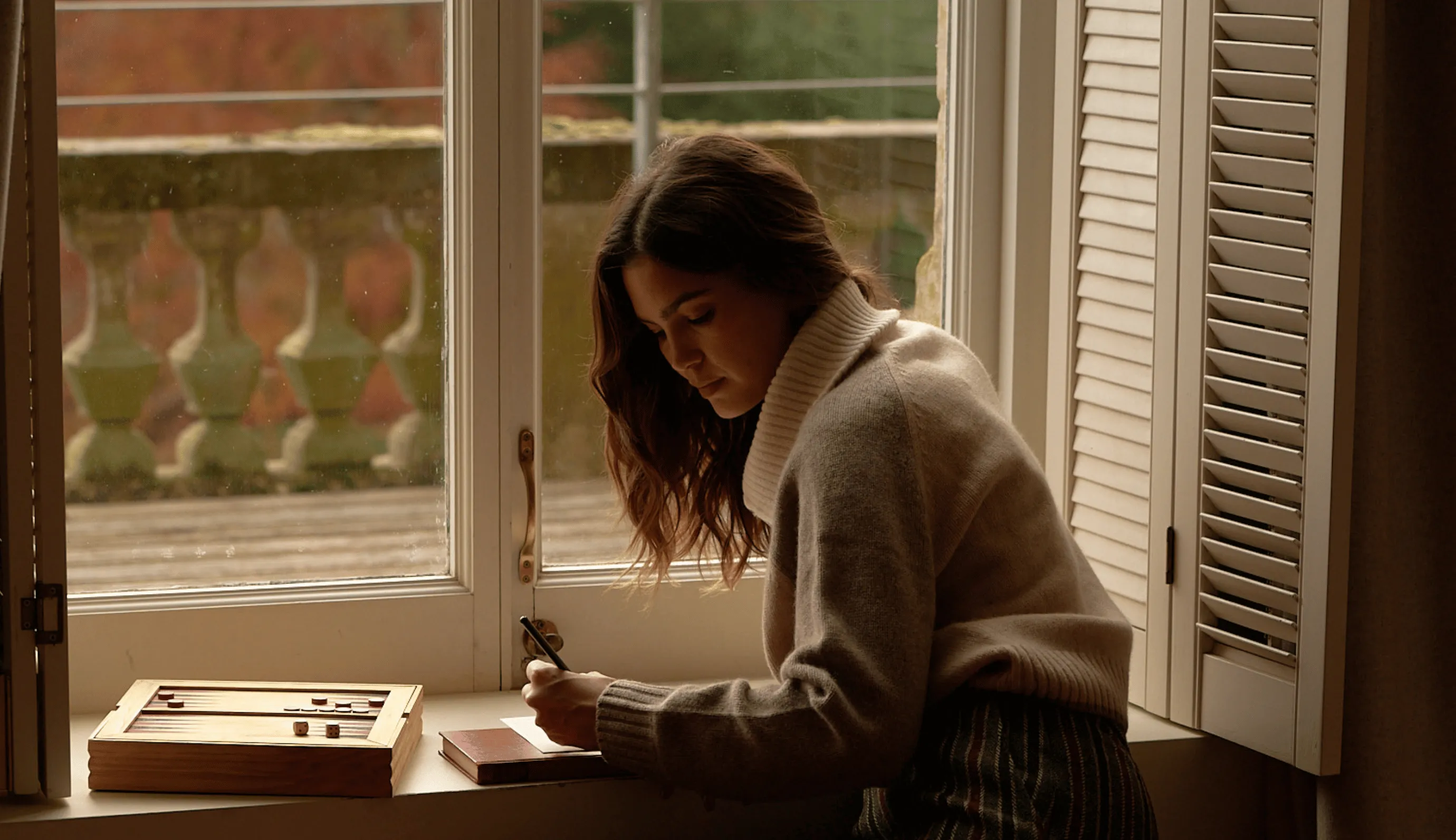 Woman in a cozy sweater writing on a notepad while sitting at a windowsill with a wooden game board nearby.