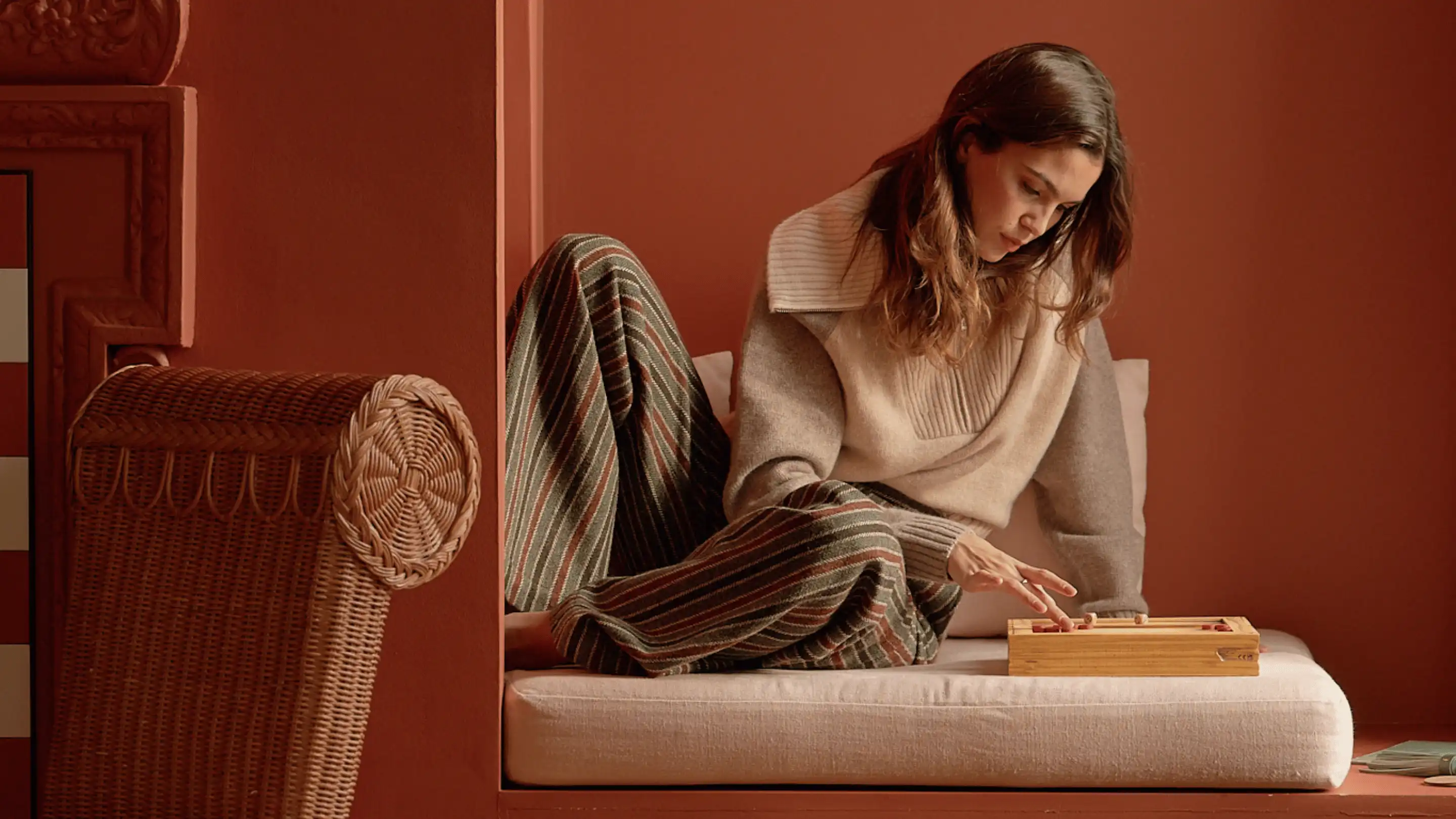 Woman wearing cozy sweater and striped pants sitting cross-legged on a cushioned window seat playing a wooden board game.