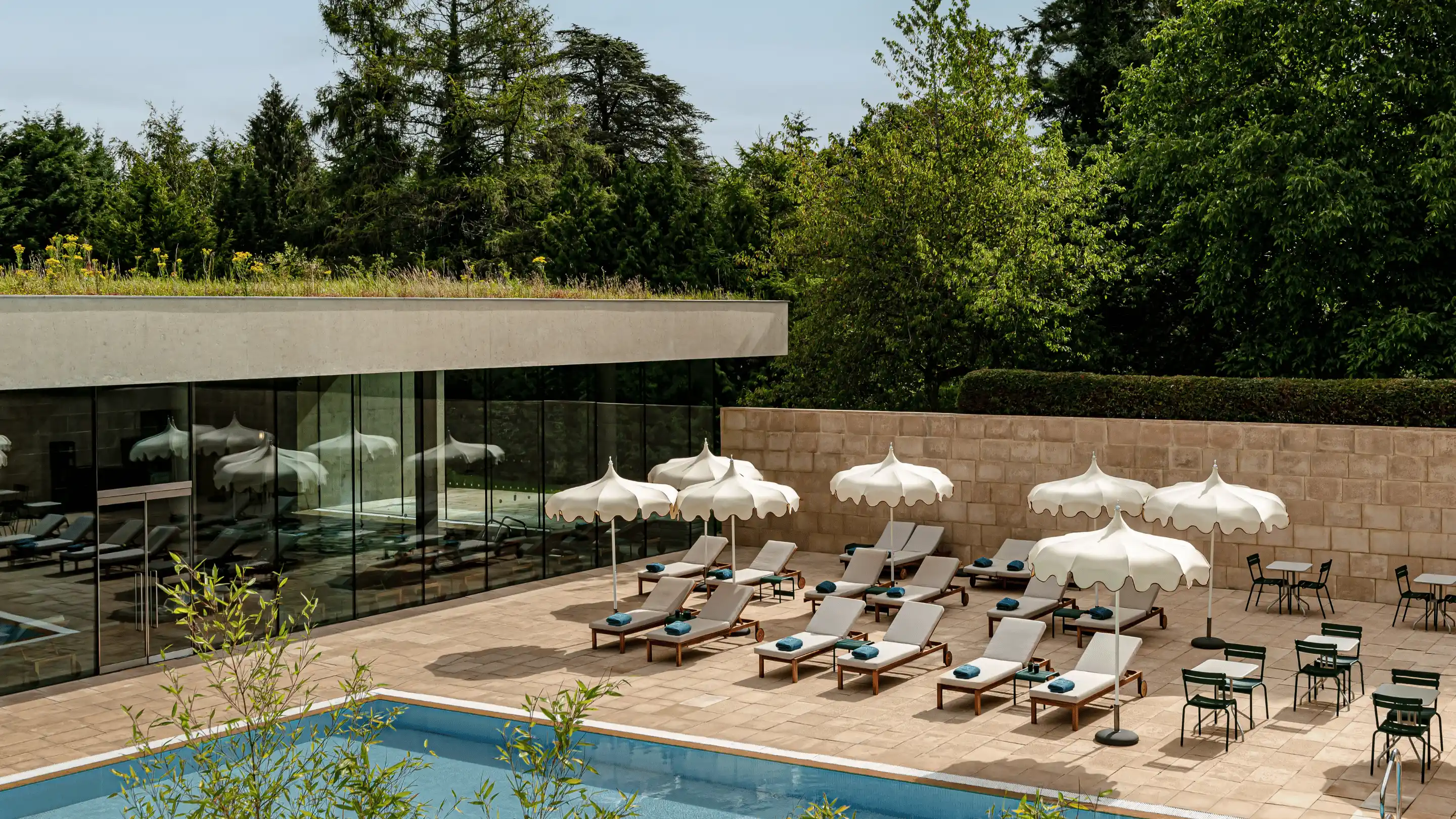 Outdoor hotel pool area with lounge chairs, white umbrellas, and small tables surrounded by trees and glass walls.