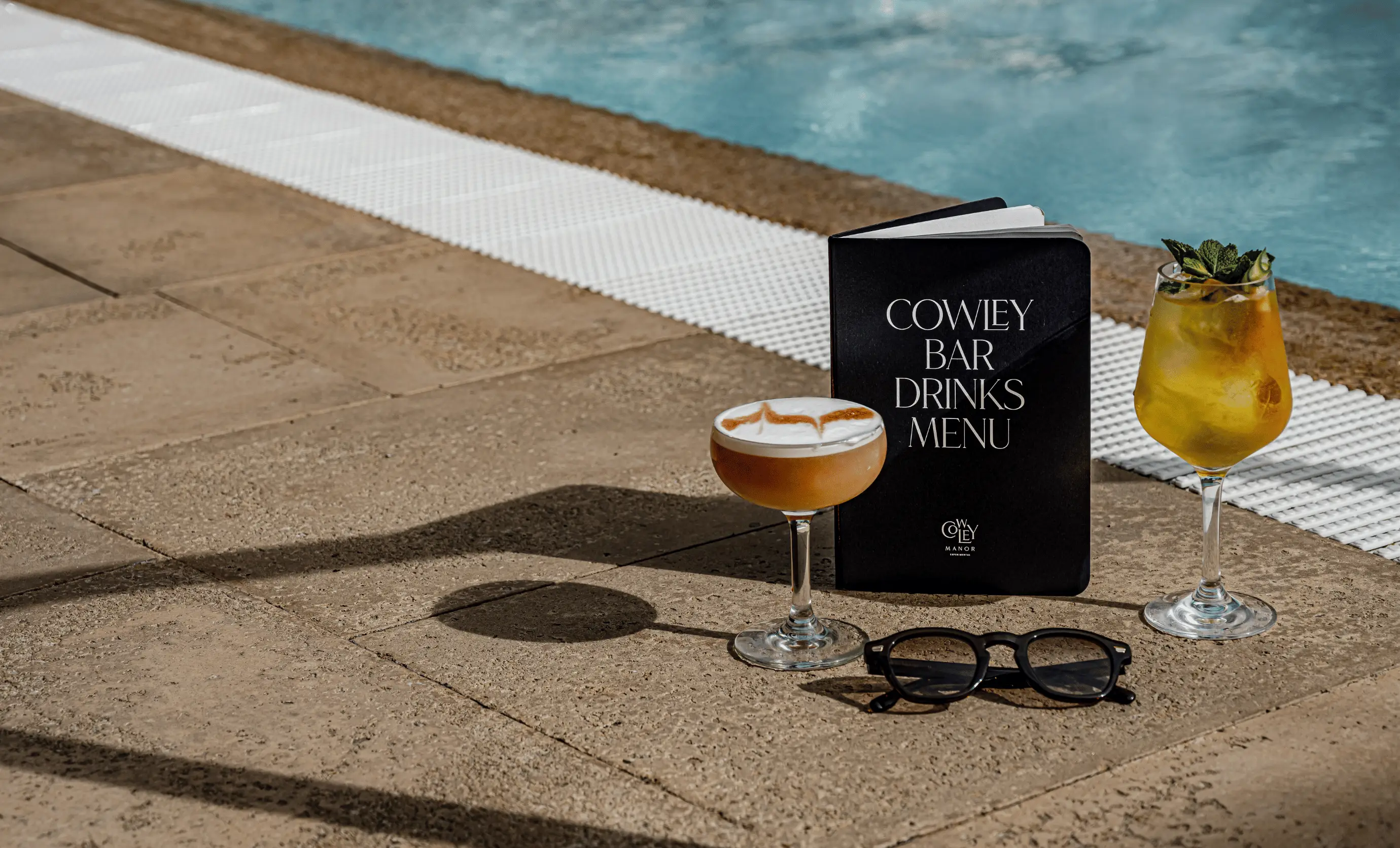 Two cocktails, a pair of black sunglasses, and a Cowley Bar drinks menu placed on a pool deck.