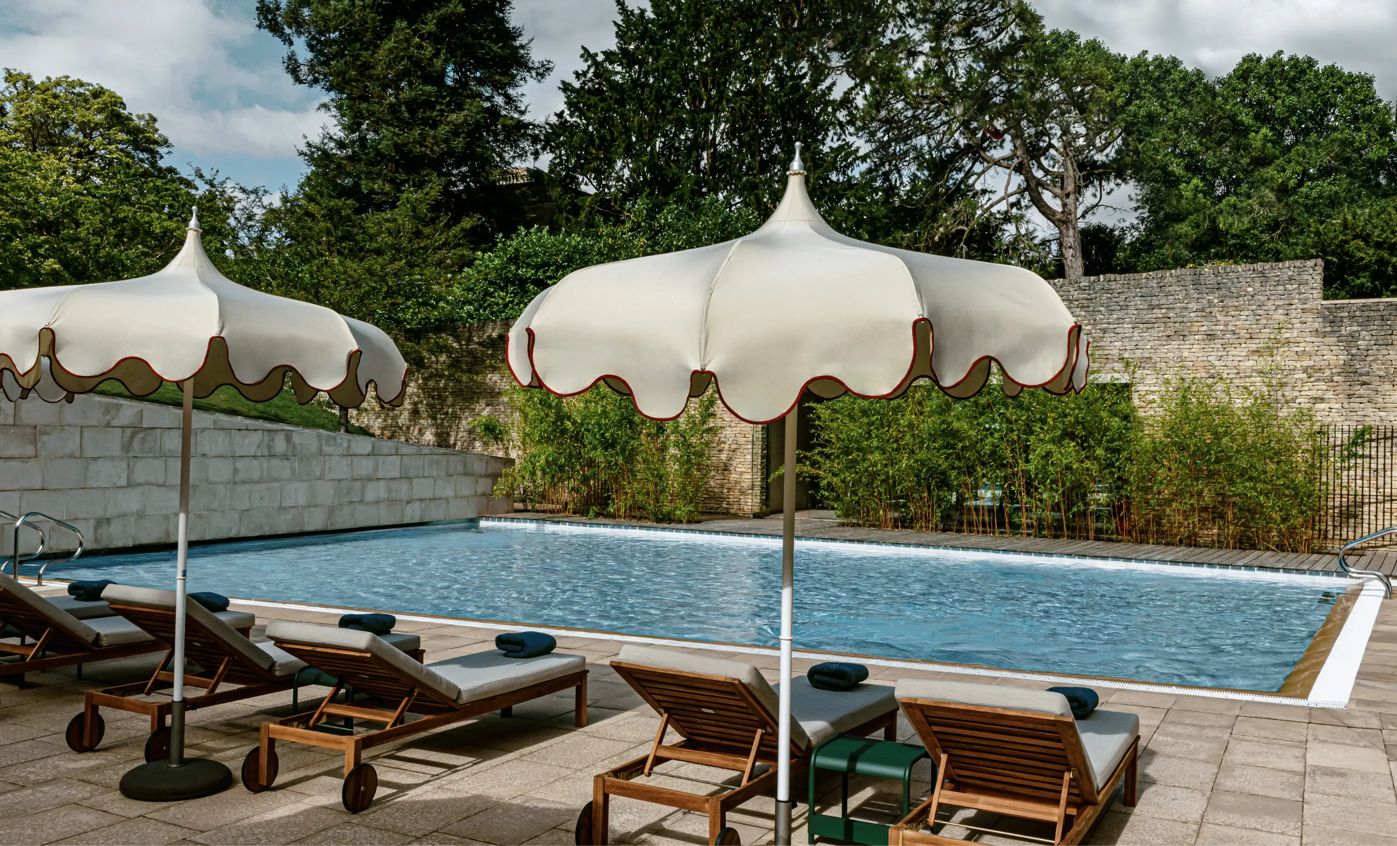 Outdoor pool area with wooden lounge chairs topped with rolled towels and white scalloped umbrellas, surrounded by greenery and stone walls.