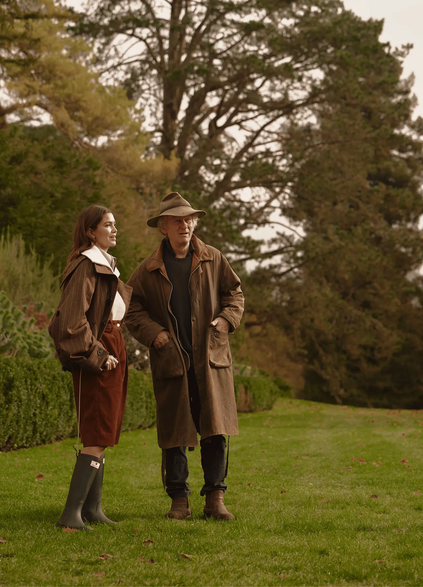 A man and a woman standing on grass with trees in the background, both wearing brown coats and looking ahead.