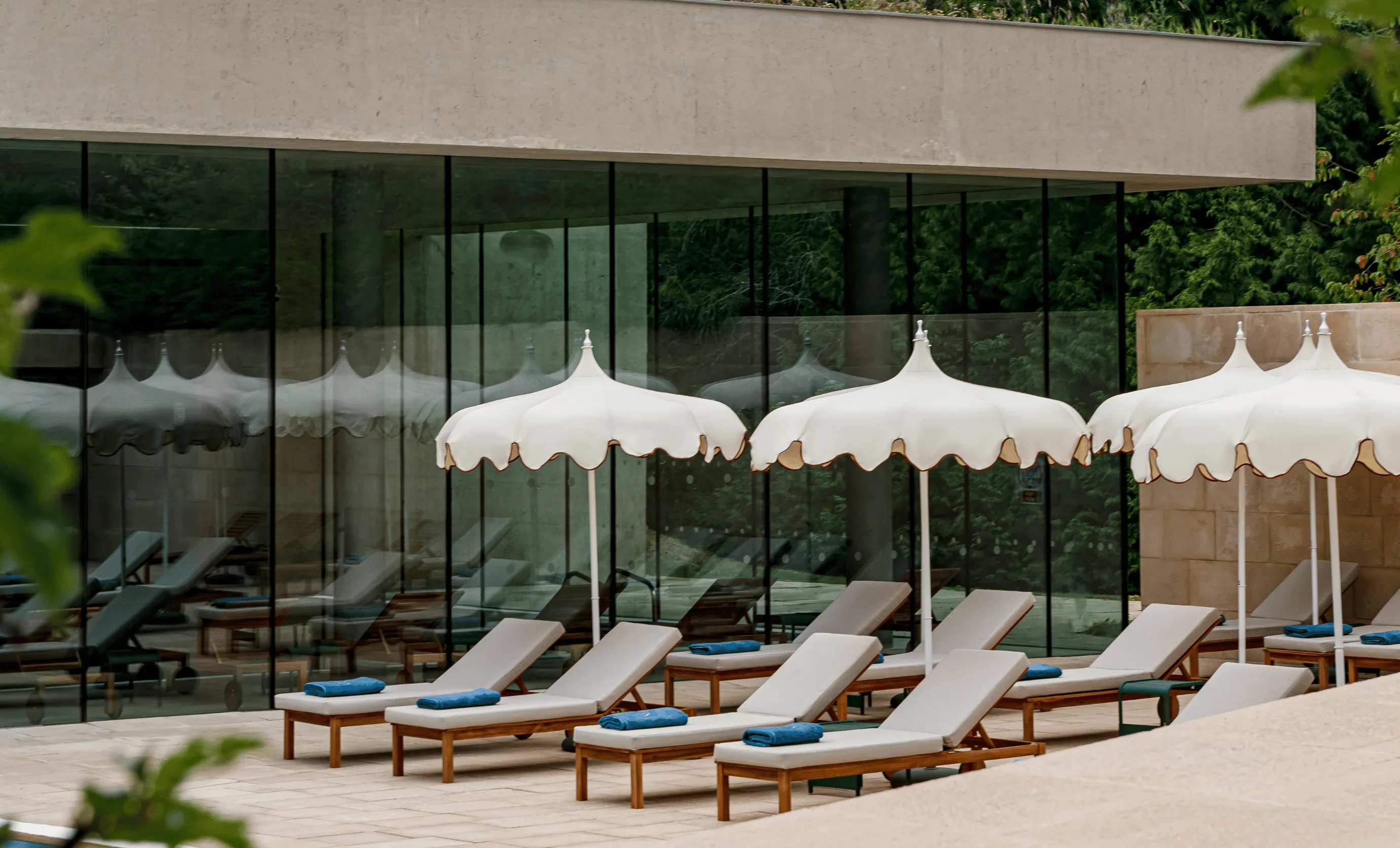 Outdoor poolside area with several wooden lounge chairs topped with white cushions and blue towels, shaded by white scalloped umbrellas, next to a modern glass building.