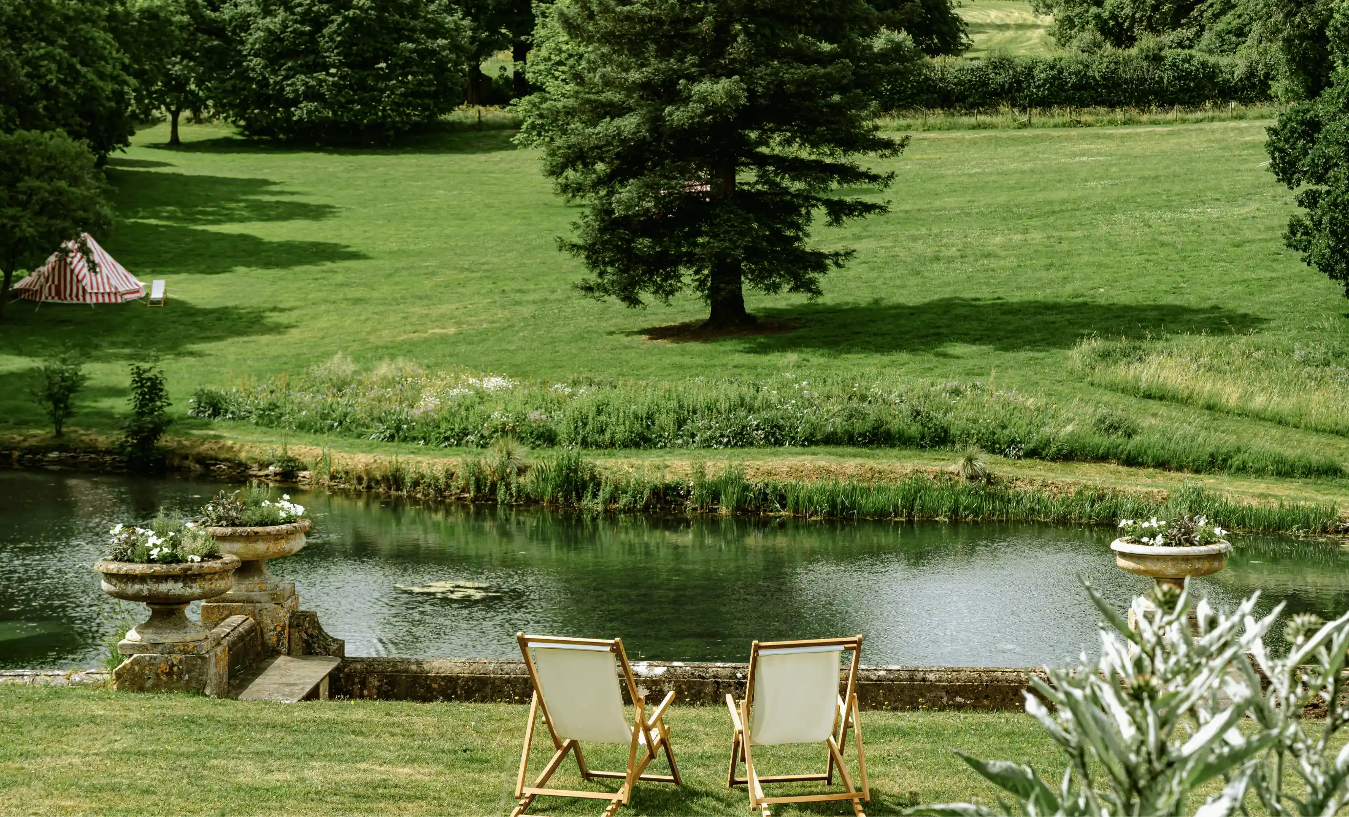 Two wooden deck chairs facing a calm river with grassy banks and a large tree casting shadows.