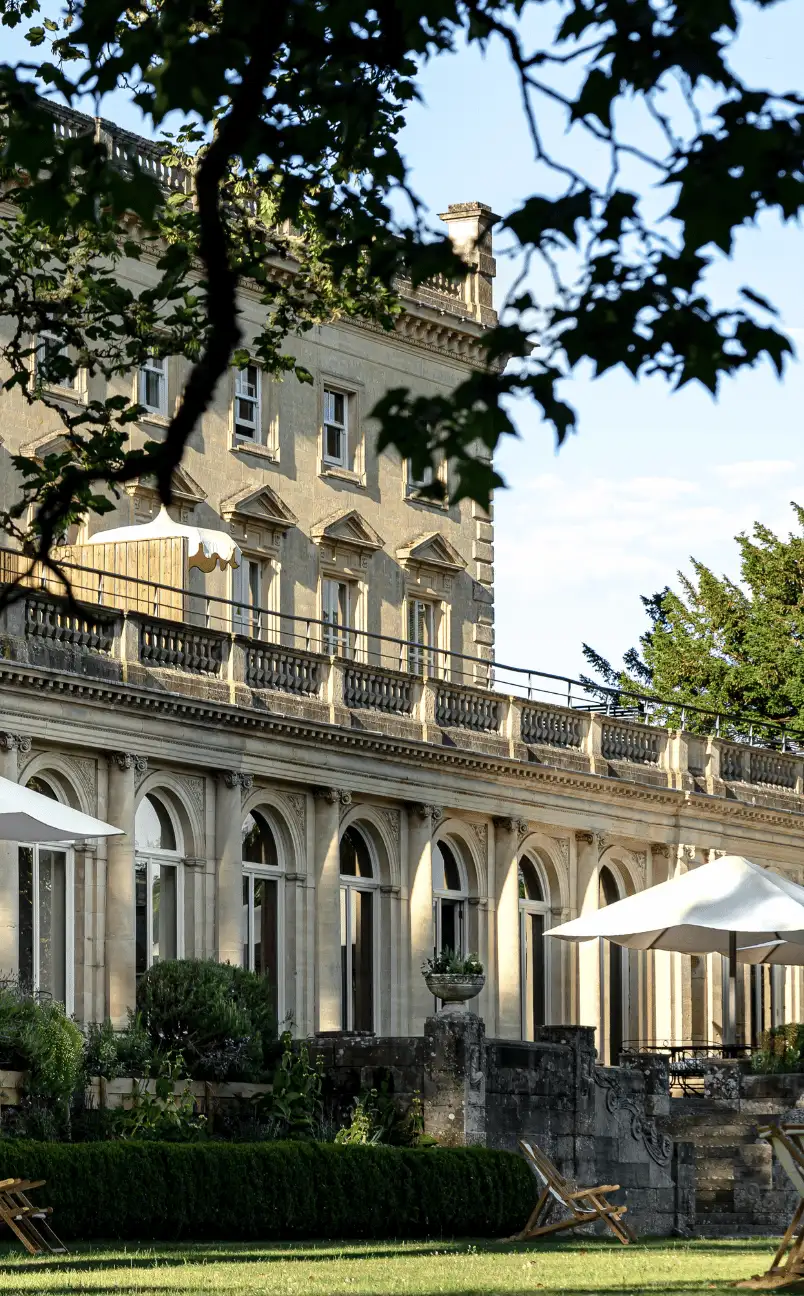 Historic stone building with arched windows and outdoor seating shaded by umbrellas, viewed through tree branches.