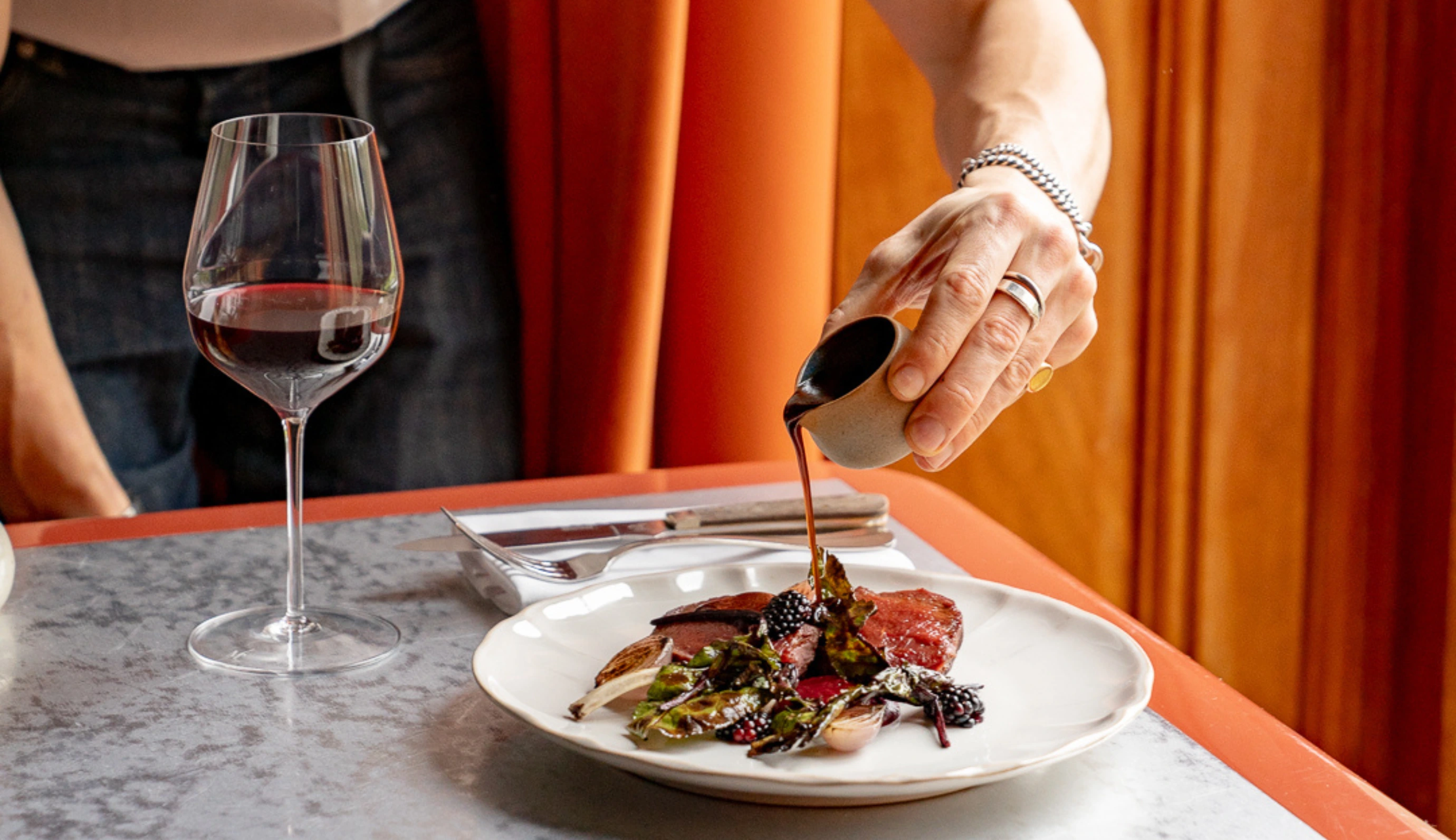 Hand pouring sauce over a gourmet dish with meat, greens, and blackberries on a white plate next to a glass of red wine.