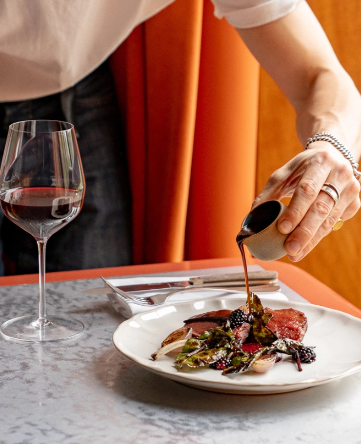 Hand pouring sauce from a small cup onto a plated steak dish with greens and blackberries next to a glass of red wine on a marble table.