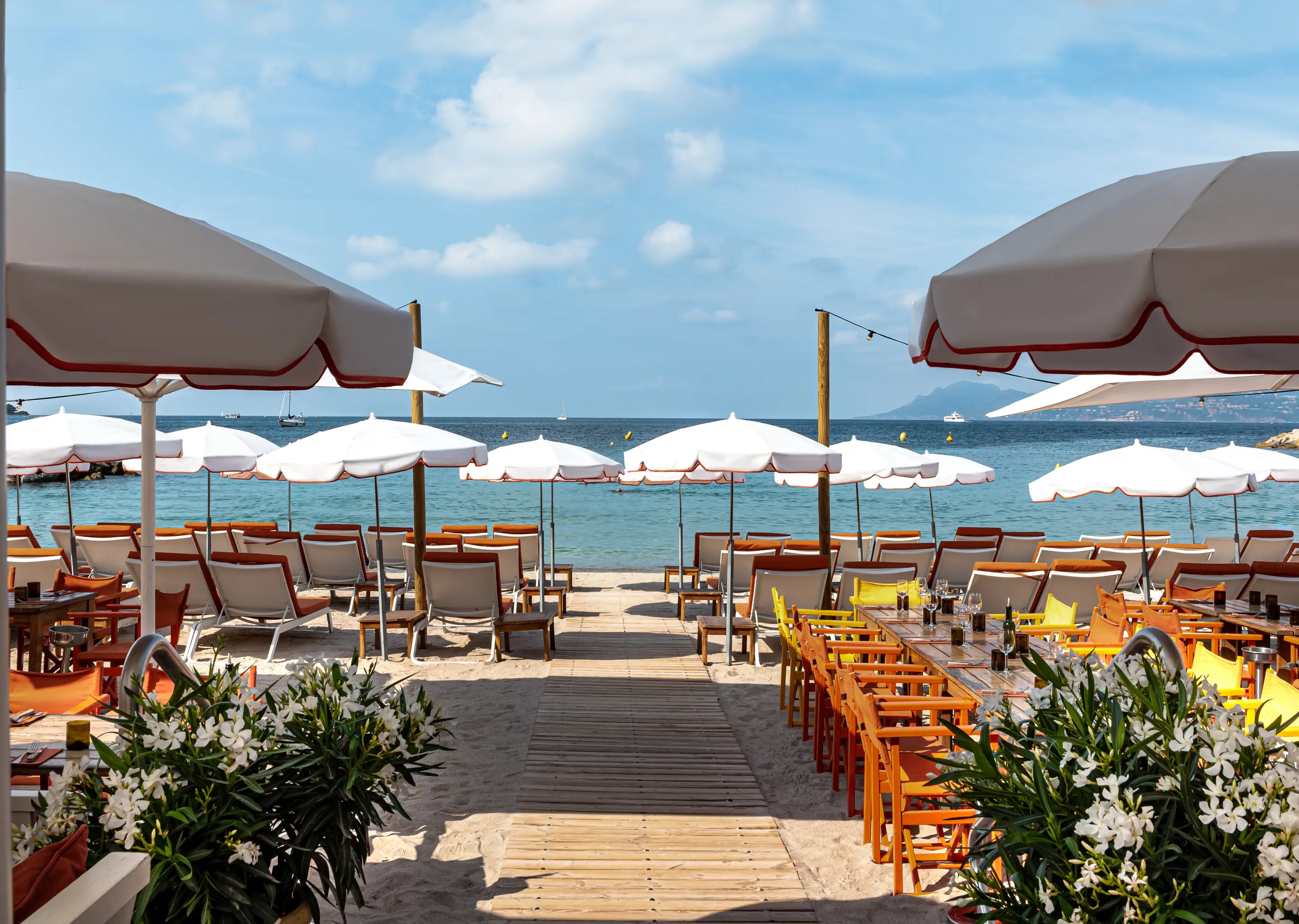 Beachside dining area with orange and yellow chairs, white umbrellas, and a wooden walkway leading to the sea under a partly cloudy sky.