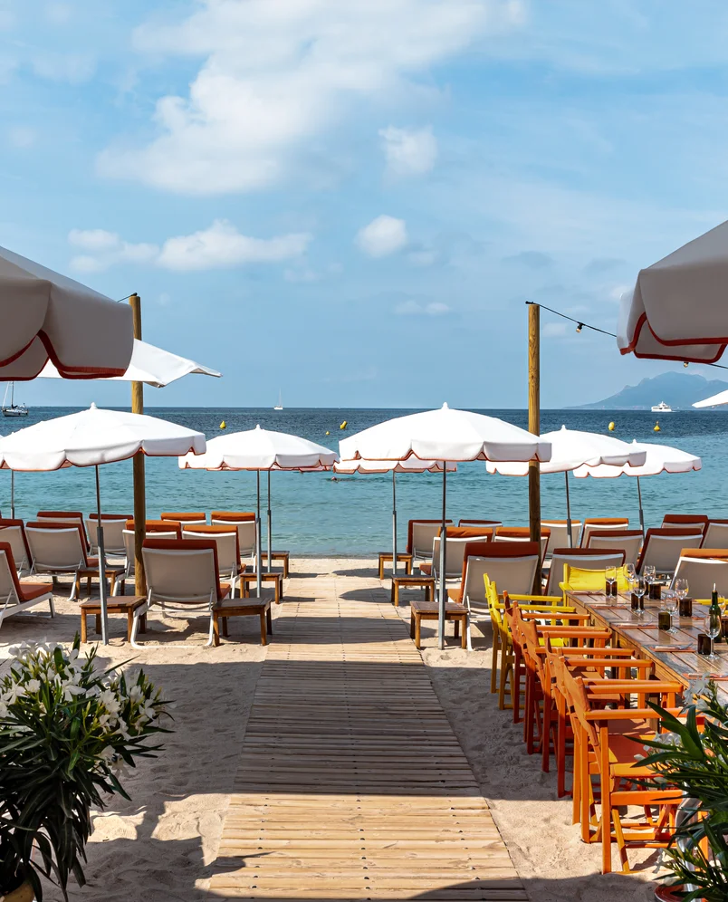 Beachfront with wooden walkway leading to rows of white umbrellas and sun loungers facing calm sea under blue sky.