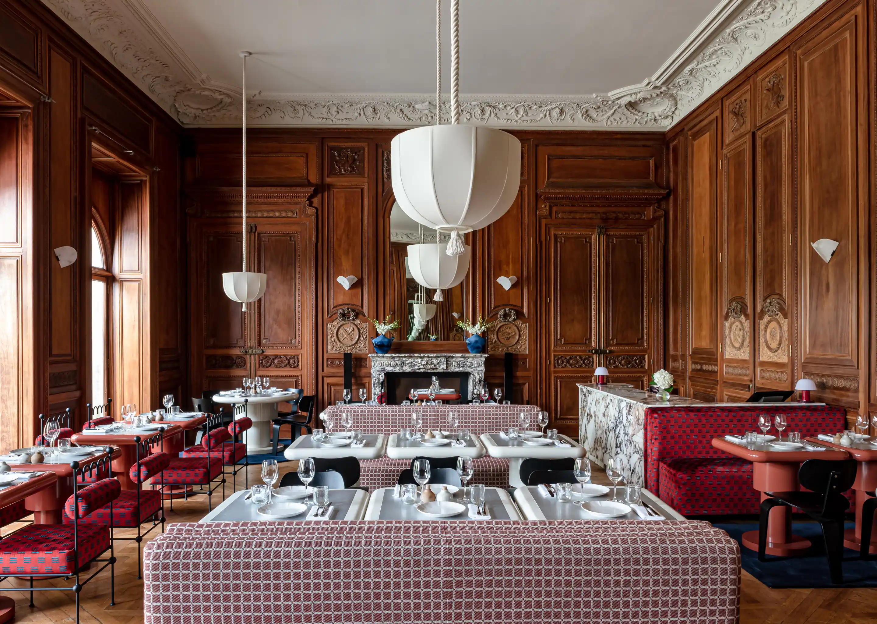 Elegant restaurant interior with wooden paneled walls, patterned upholstered seating, white tables set with plates and glassware, and large white pendant lights.