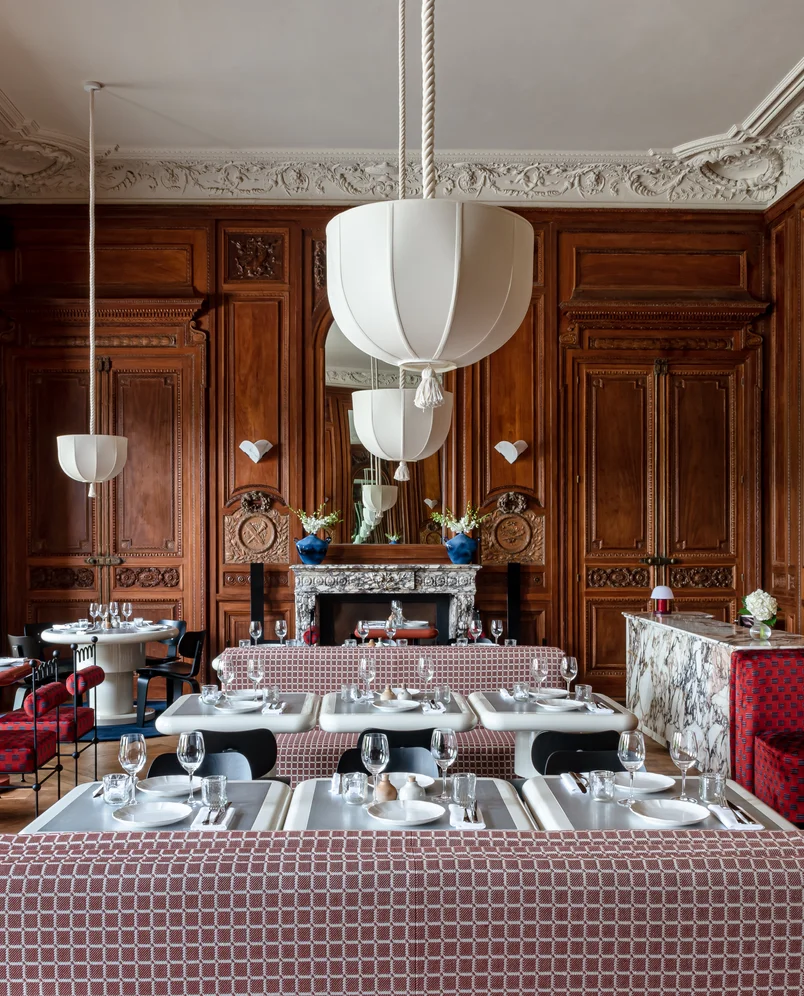 Elegant restaurant interior with ornate wooden wall panels, white pendant lights, marble fireplace, and tables set with glassware and plates.