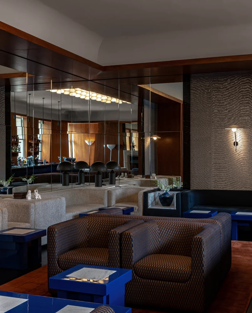 Modern lounge with patterned armchairs, cream sofas, blue tables, wall mirrors, and warm ambient lighting.