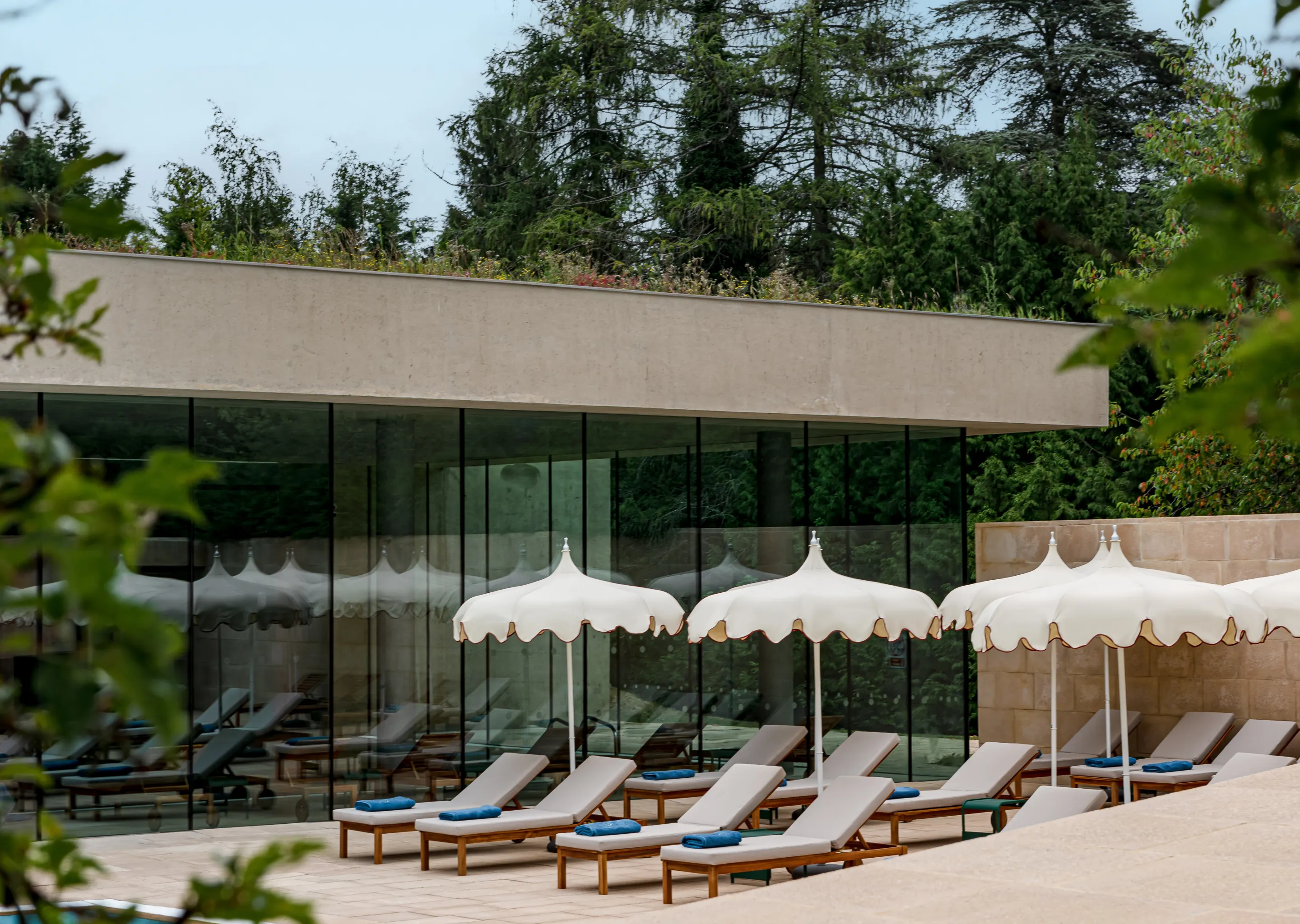 Row of cushioned lounge chairs with blue towels under white parasols beside a modern glass building, surrounded by greenery.