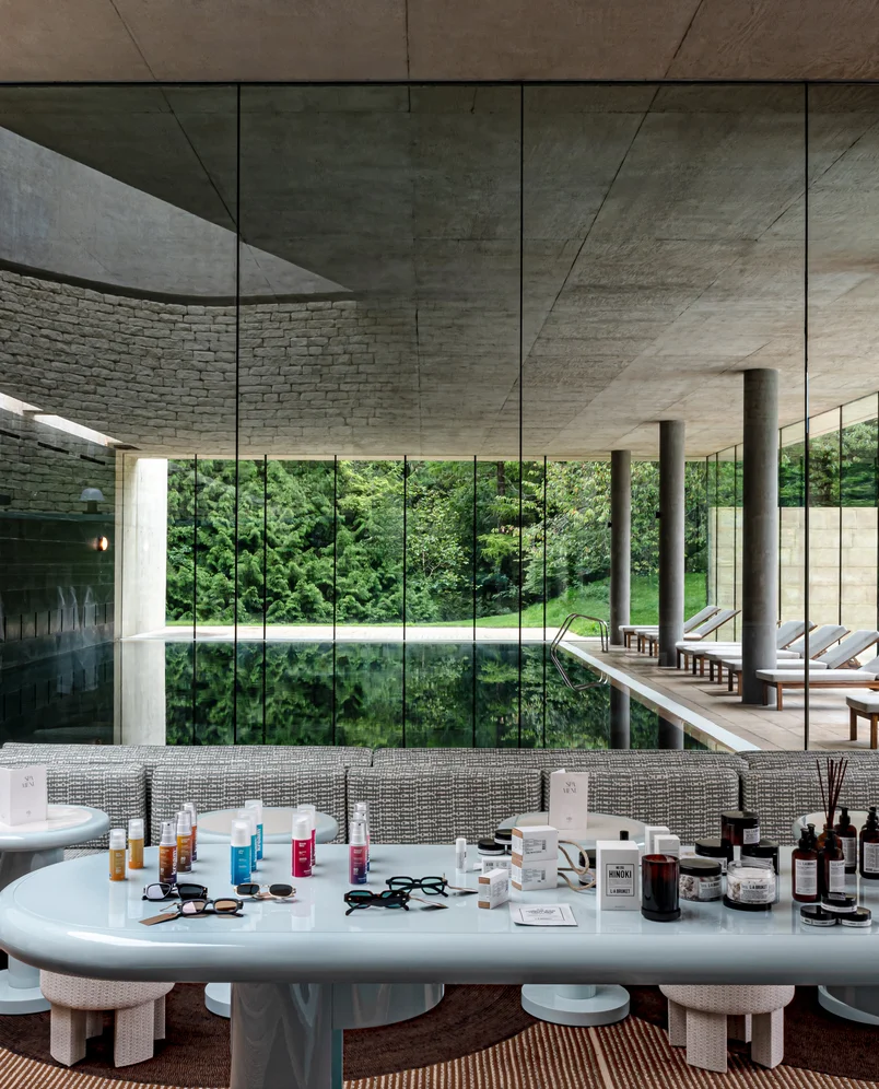 Indoor pool area with glass walls overlooking lush greenery, a modern stone ceiling, lounge chairs on the right, and a table in the foreground displaying various skincare products and glasses.