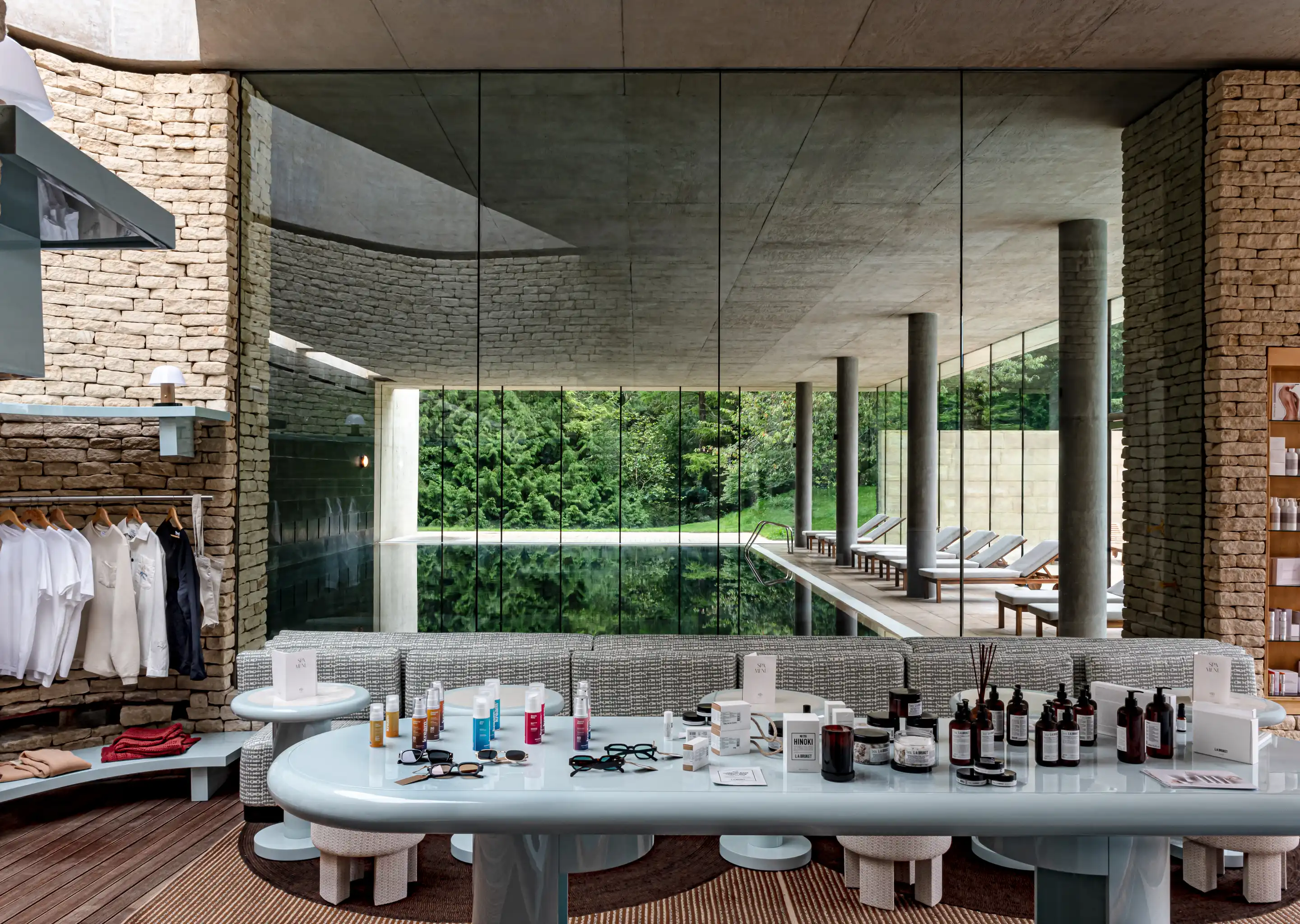Modern indoor pool area with glass walls, lounge chairs, and a table displaying skincare products and sunglasses in front.