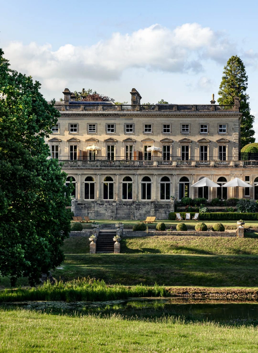 Large elegant manor house with multiple windows and outdoor umbrellas, surrounded by manicured gardens and trees.