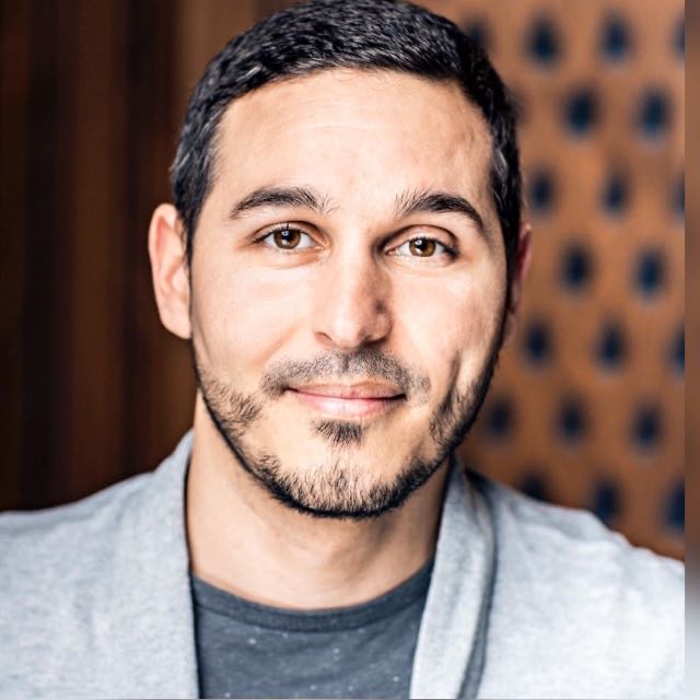 Close-up portrait of a man with short dark hair, brown eyes, and light facial hair, wearing a gray shirt and light jacket, smiling gently against a blurred background.