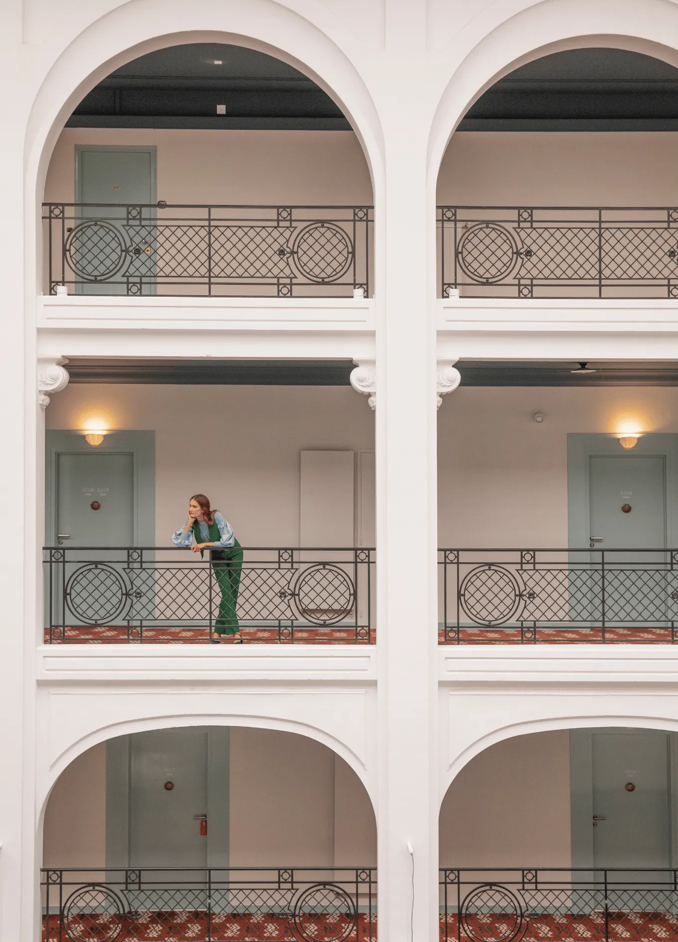 Woman in green jumpsuit leaning on the railing of a hotel corridor with arched openings and patterned carpet.