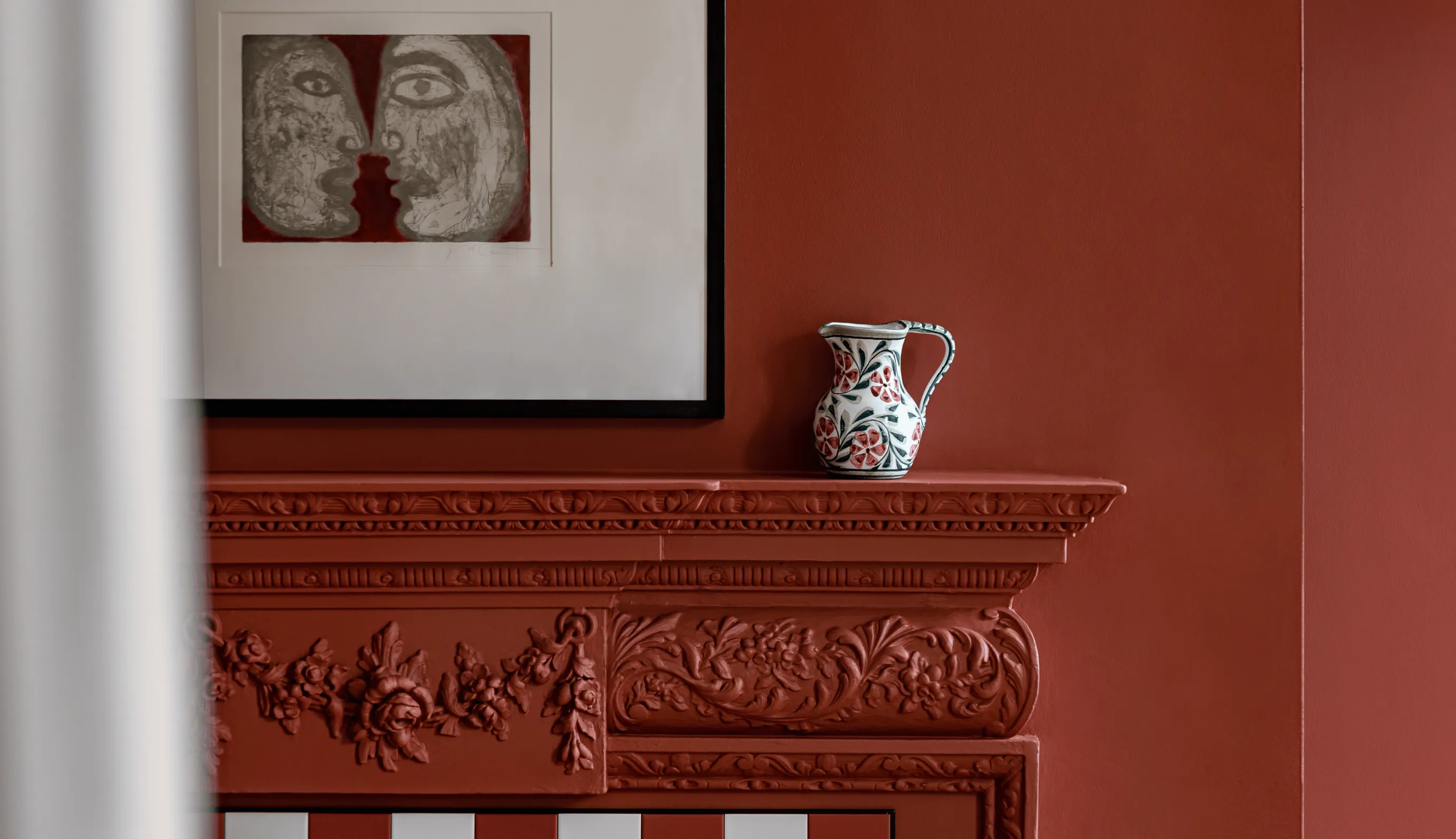 Decorative ceramic pitcher with floral patterns on a red, intricately carved mantel beneath a framed artwork of two abstract faces.