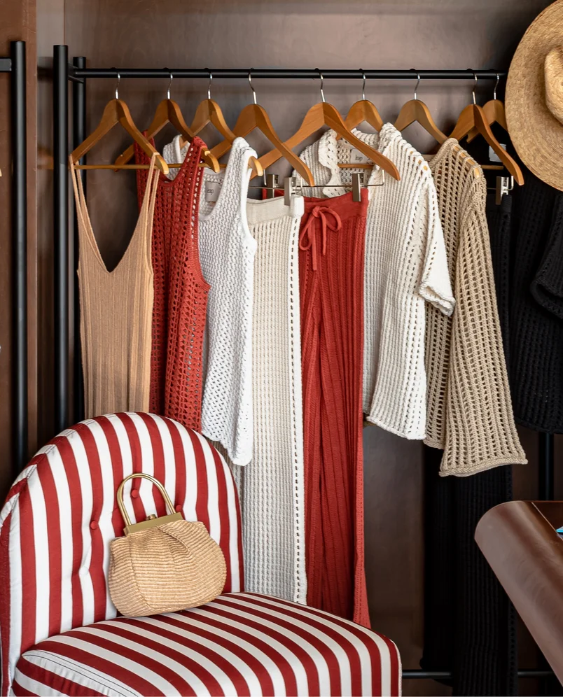Clothing rack with knitted dresses and sweaters in neutral and rust colors, next to a red and white striped chair with a woven handbag.