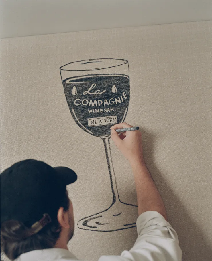 Person drawing a logo of a wine glass with the text 'La Compagnie Wine Bar New York' on a beige canvas.