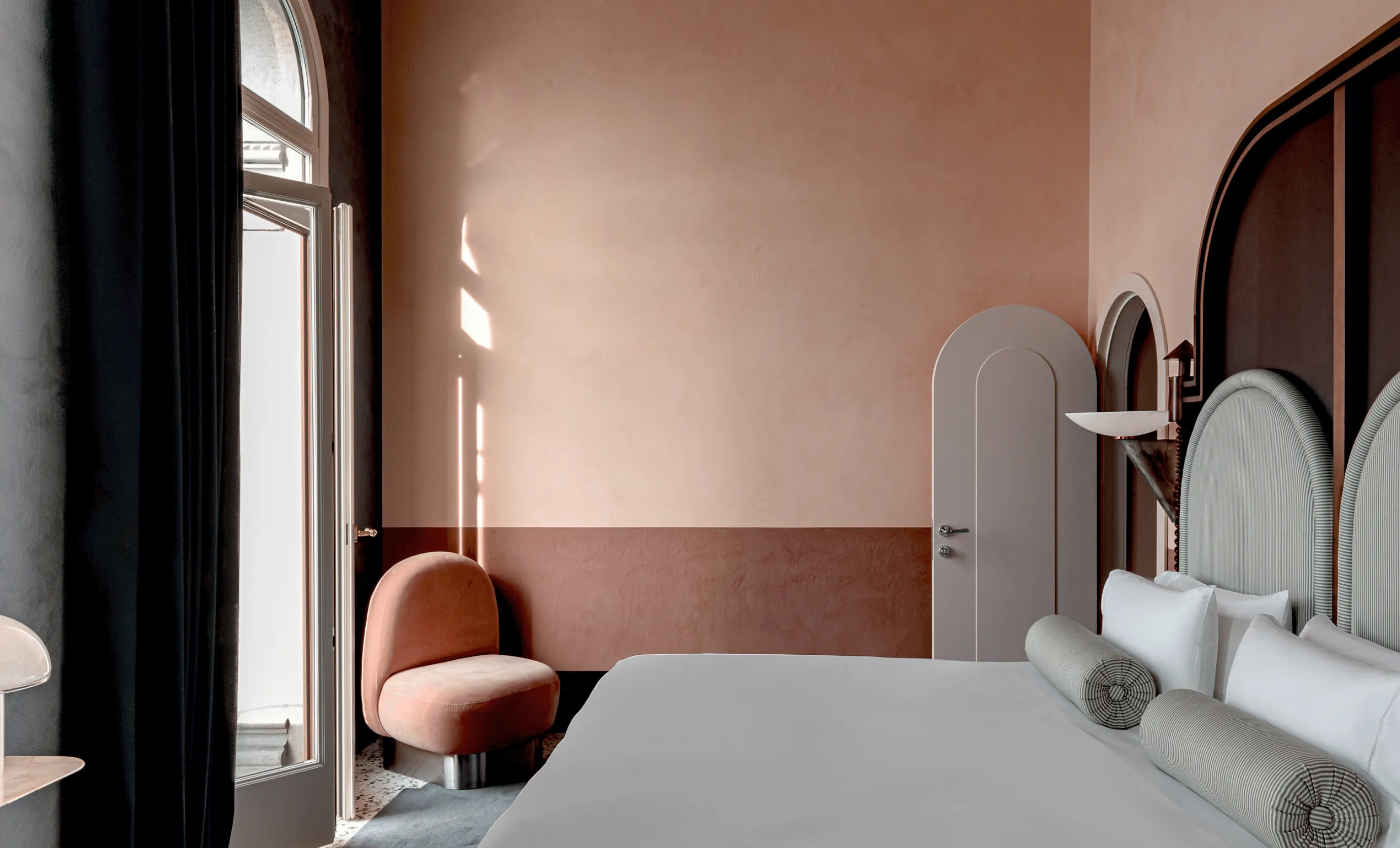 Modern hotel bedroom with muted pink walls, a large bed with round gray pillows, and a peach-colored rounded chair near a tall window with black curtains.