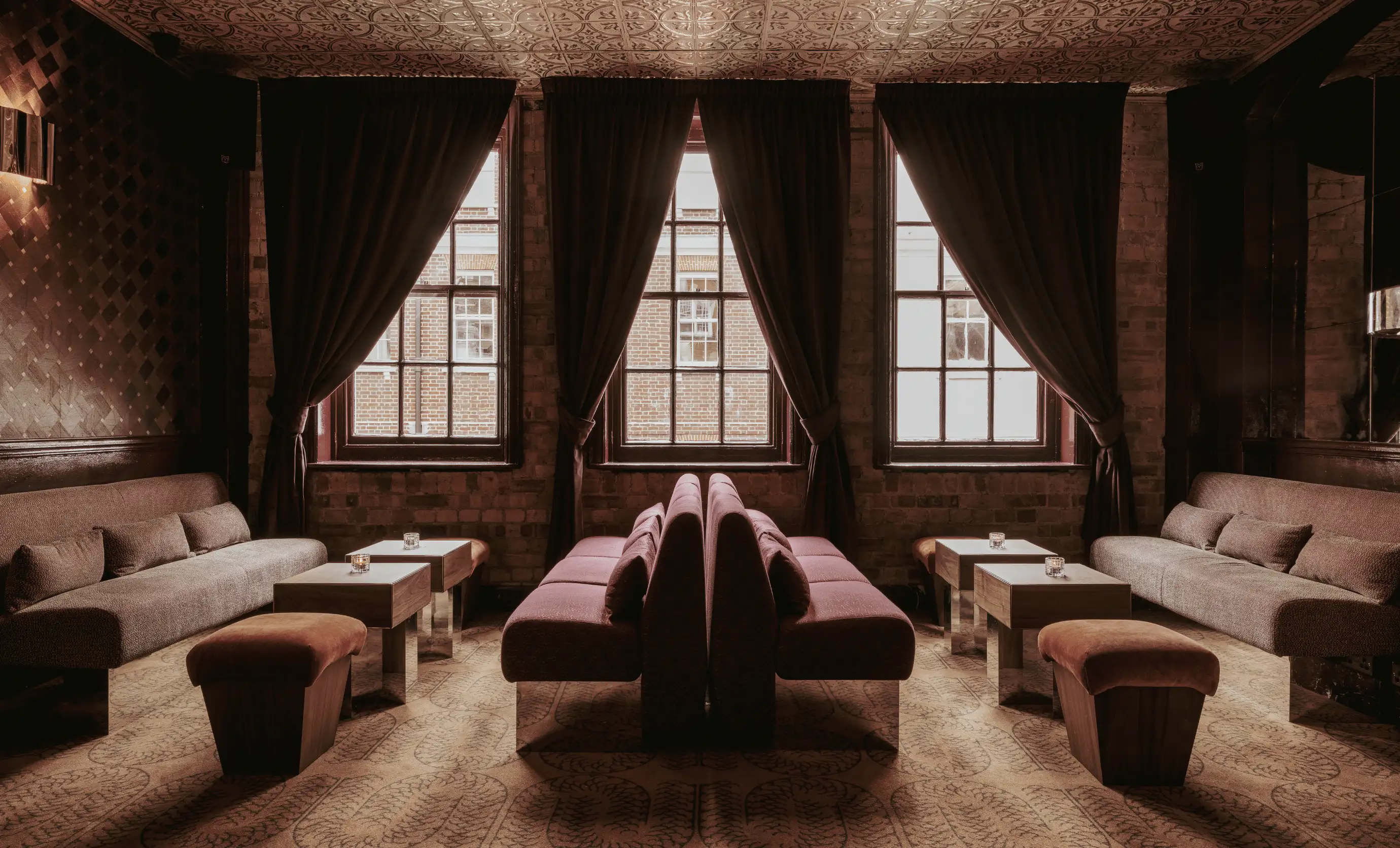 Symmetrical hotel lounge with dark purple sofas, wooden tables, beige stools, large windows with dark curtains, and patterned carpet.