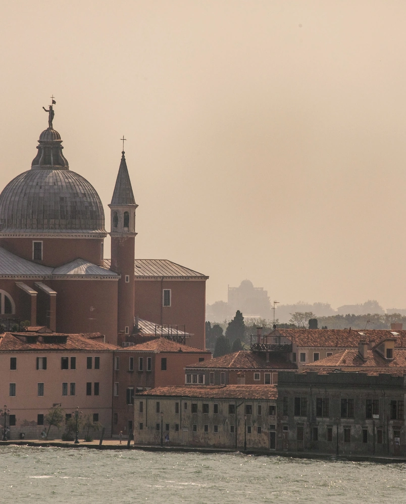 Historic buildings with a large dome and a bell tower by a waterfront under a hazy sky.