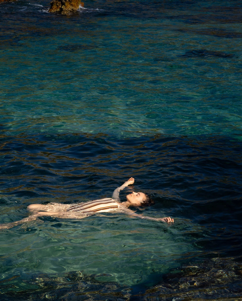 Woman in striped swimsuit floating relaxed on clear, blue ocean water near rocky shore.