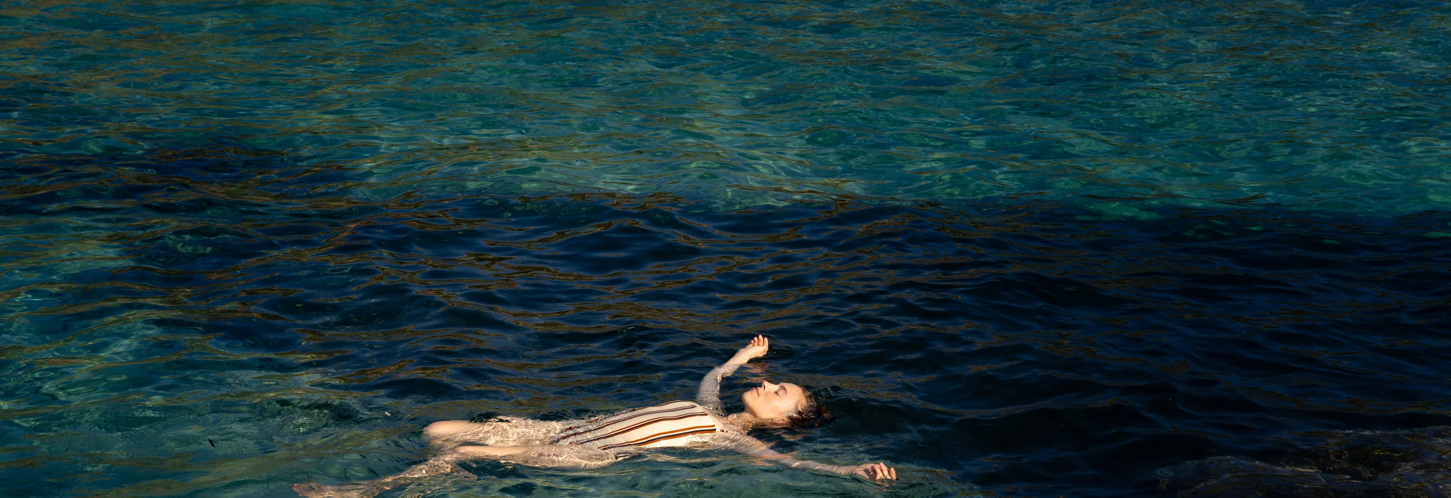 Woman in a striped swimsuit floating calmly on clear blue sea water.