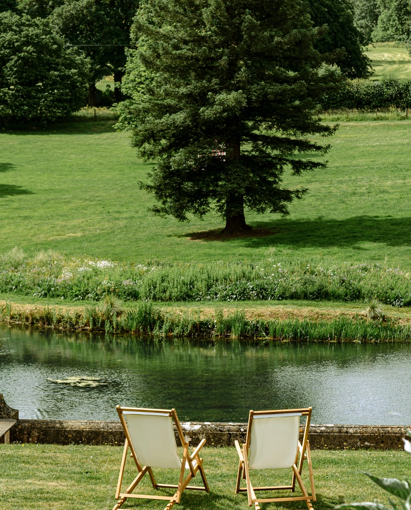 Two empty wooden deck chairs facing a calm pond with grass, trees, and lush greenery in the background.