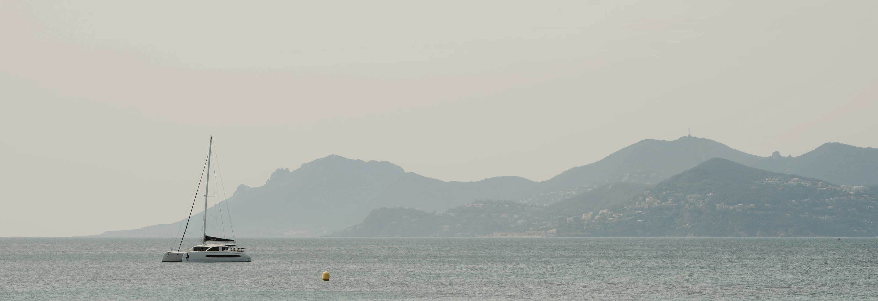 White sailboat floating on calm sea with hazy mountainous coastline in the background.