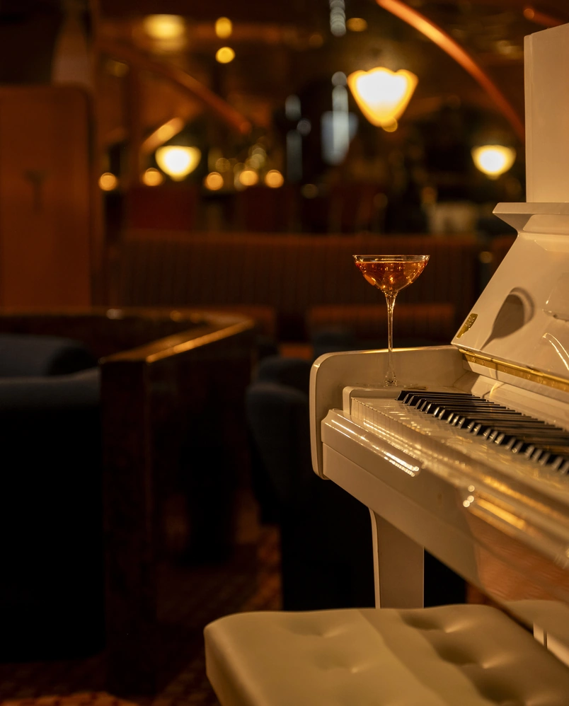 A glass of amber-colored cocktail placed on the edge of a white piano keyboard in a dimly lit lounge.
