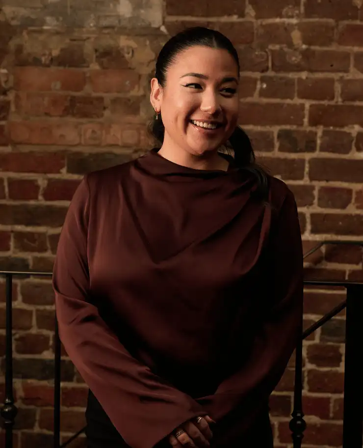 Smiling woman with dark hair tied back wearing a brown long-sleeve top standing in front of a brick wall.
