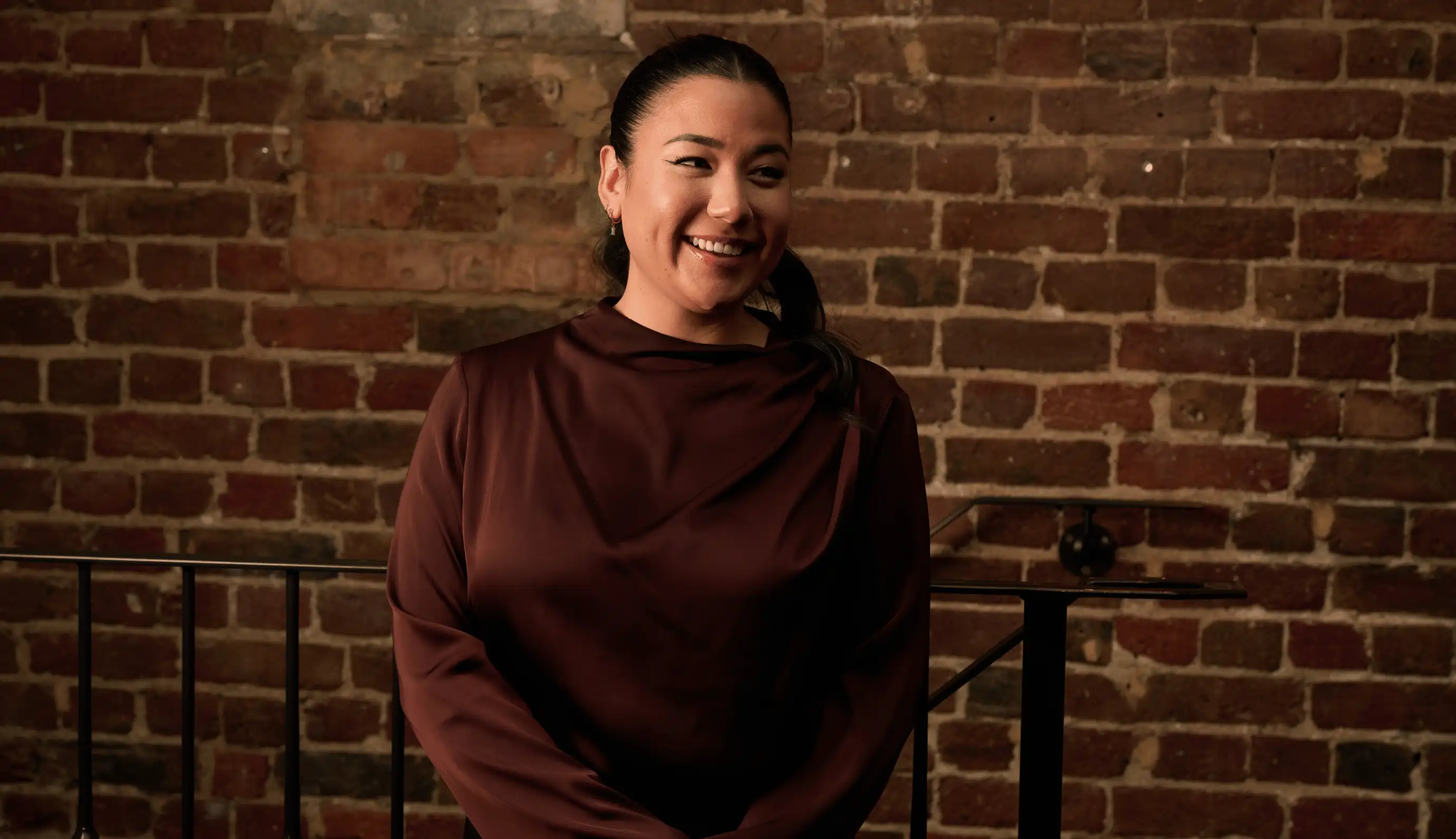 Smiling woman with dark hair in a low ponytail wearing a maroon long-sleeve top standing in front of a brick wall.