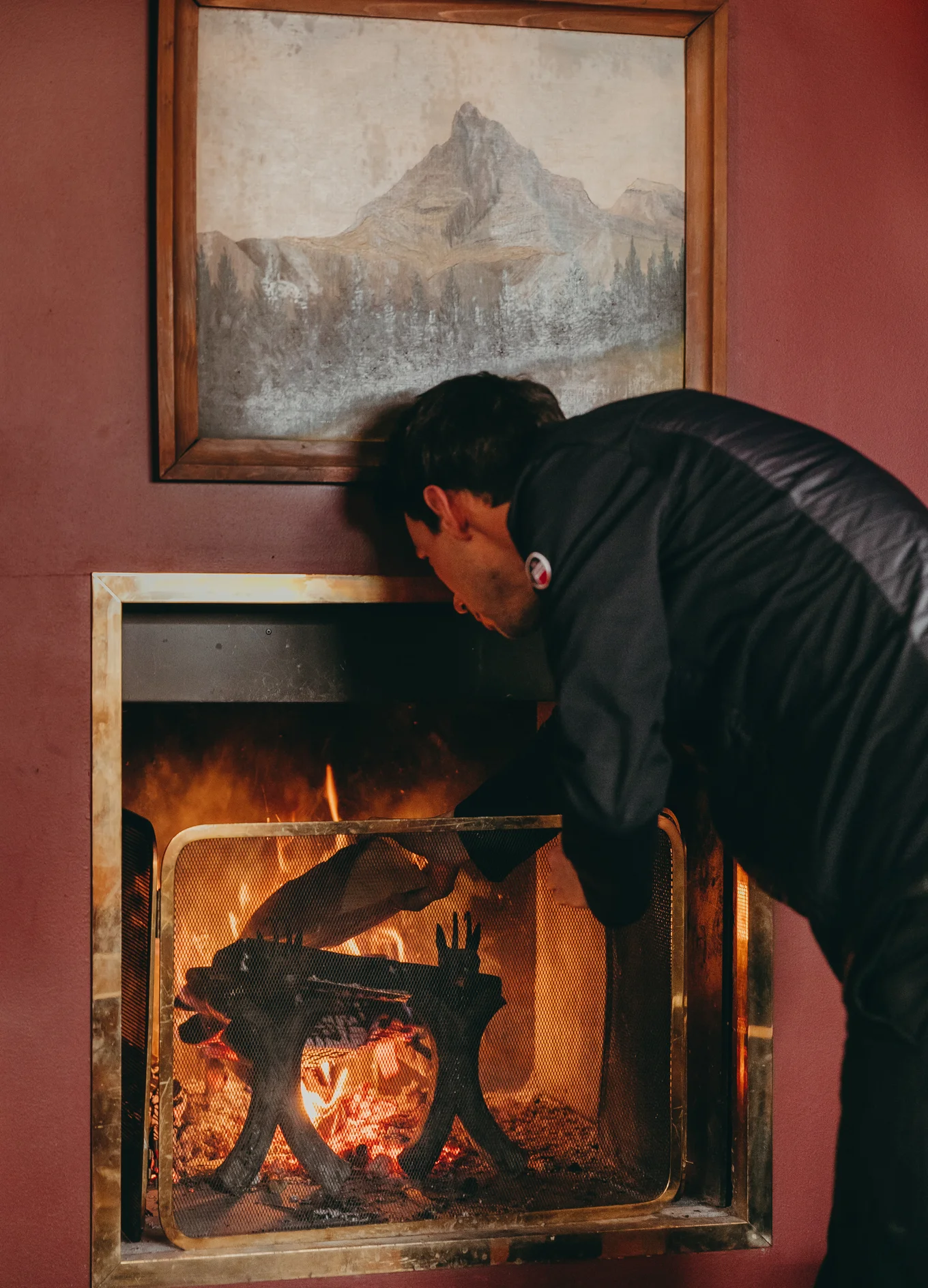 Man tending a fire in a framed indoor fireplace with a mountain painting on the wall above.