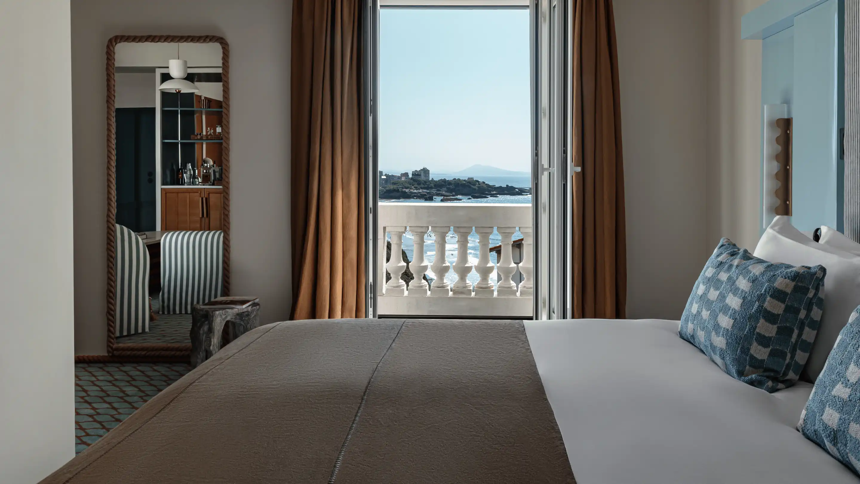 Hotel room with a bed facing an open balcony door overlooking the sea and rocky coastline.